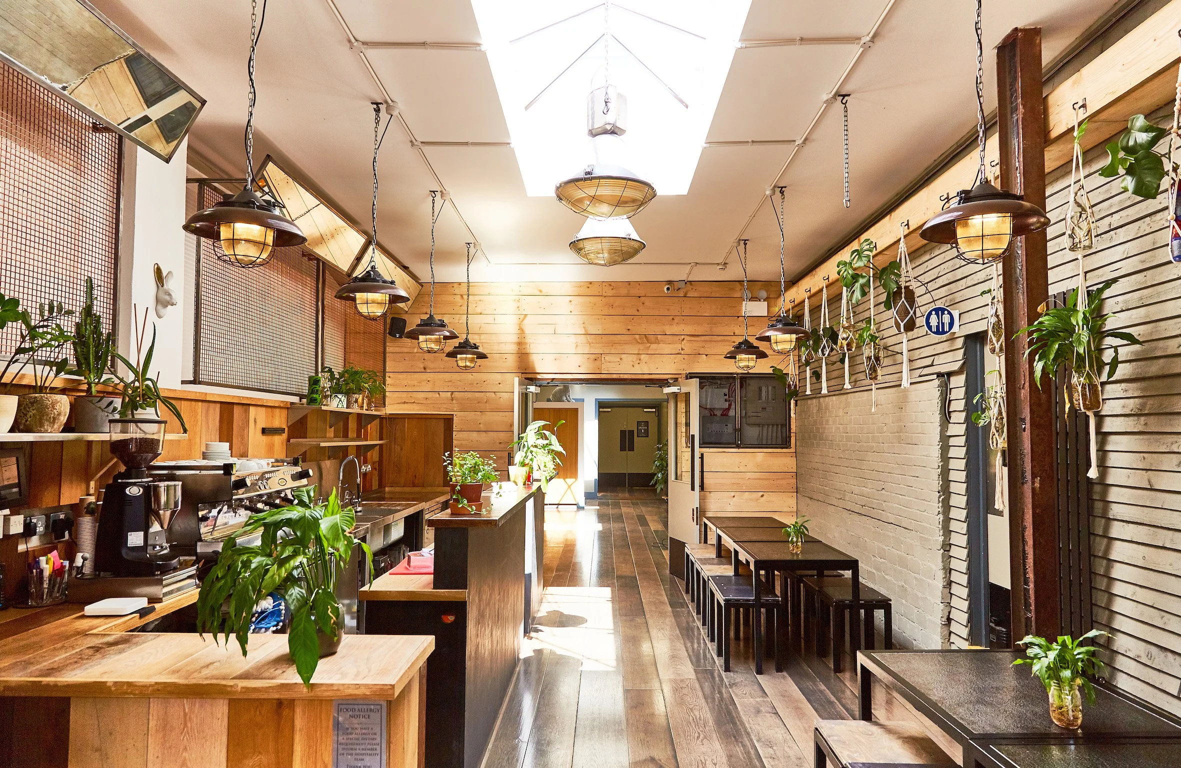 Interior of a cozy cafe or restaurant with wooden walls, tables, and plants. Hanging lights illuminate the space, and a barista station is visible on the left. There are small tables with chairs along the right wall, and a sign indicating restrooms is on the wall.