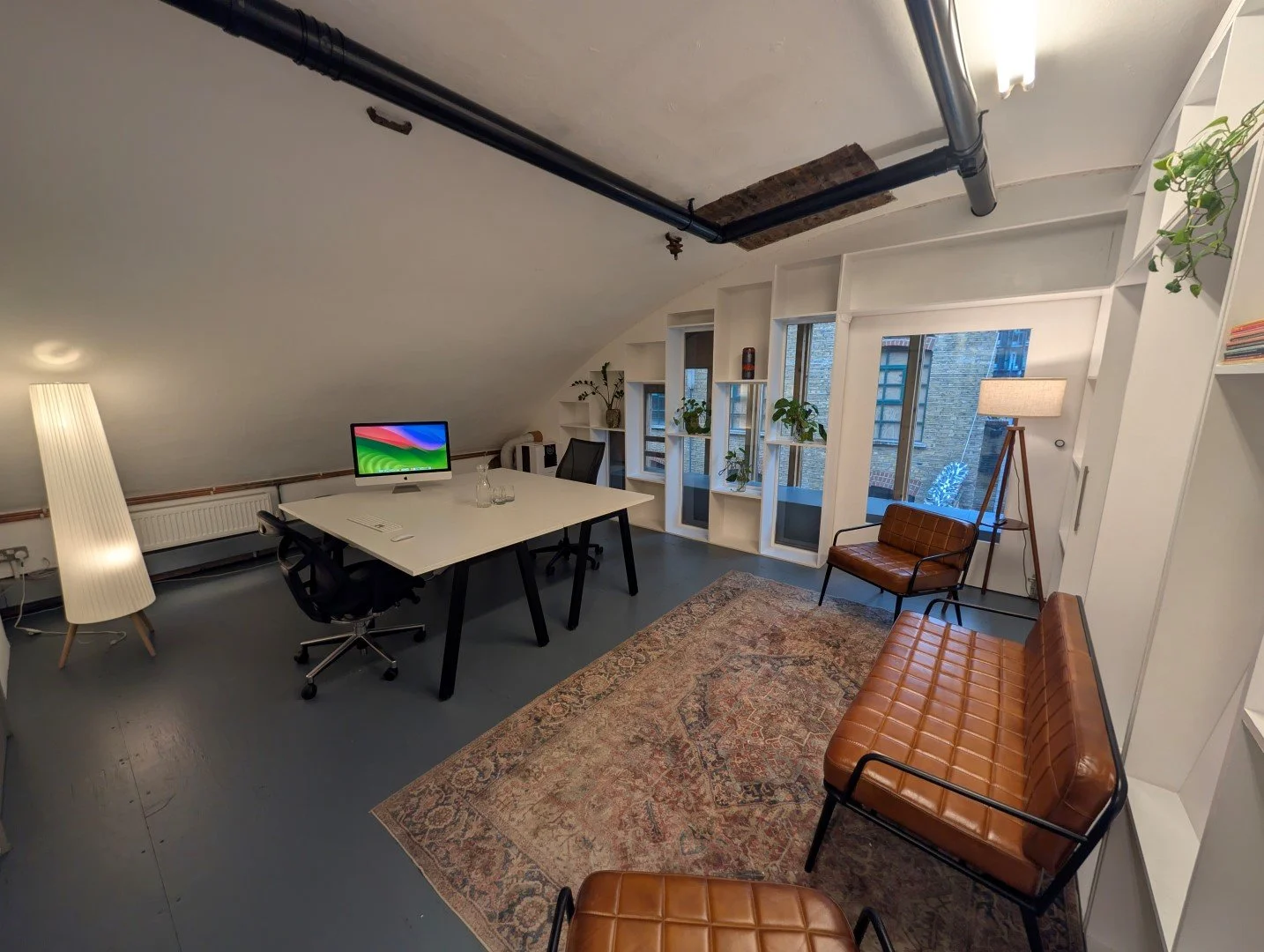 Modern office with white walls and large windows, featuring a white desk with an iMac, black office chairs, a brown leather loveseat and chair, a beige rug, a tall floor lamp, and decorative plants.