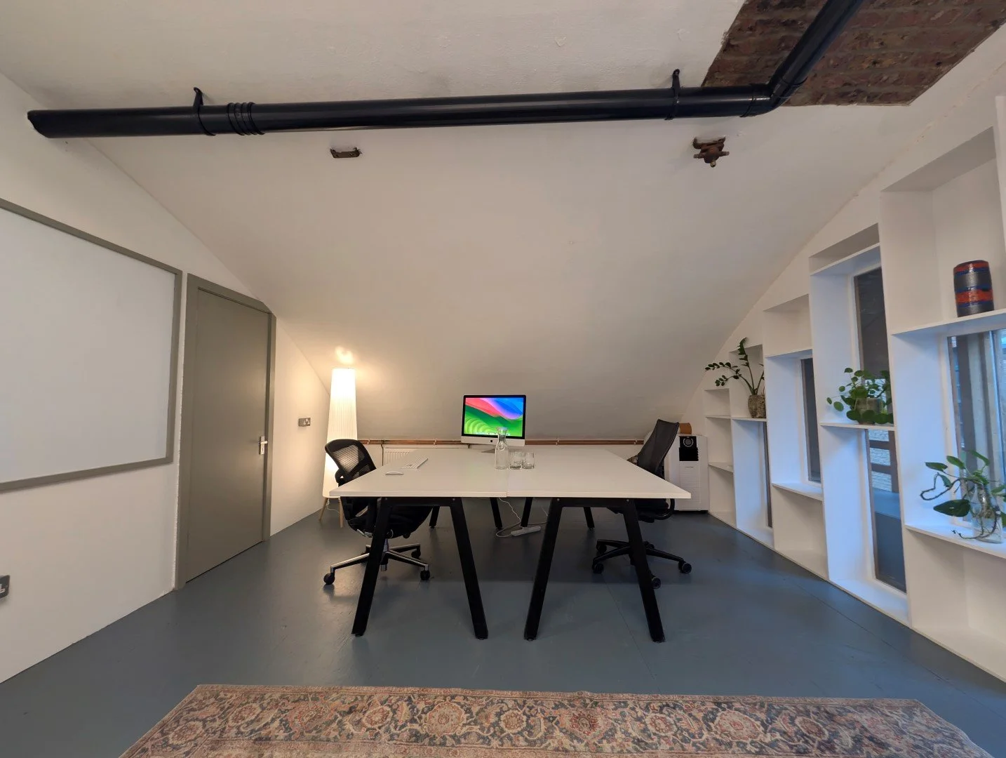 Modern office meeting room with a white table, black chairs, a computer monitor, potted plants on built-in shelves, and a whiteboard on the wall. Ceiling has exposed black pipe and brick accents.