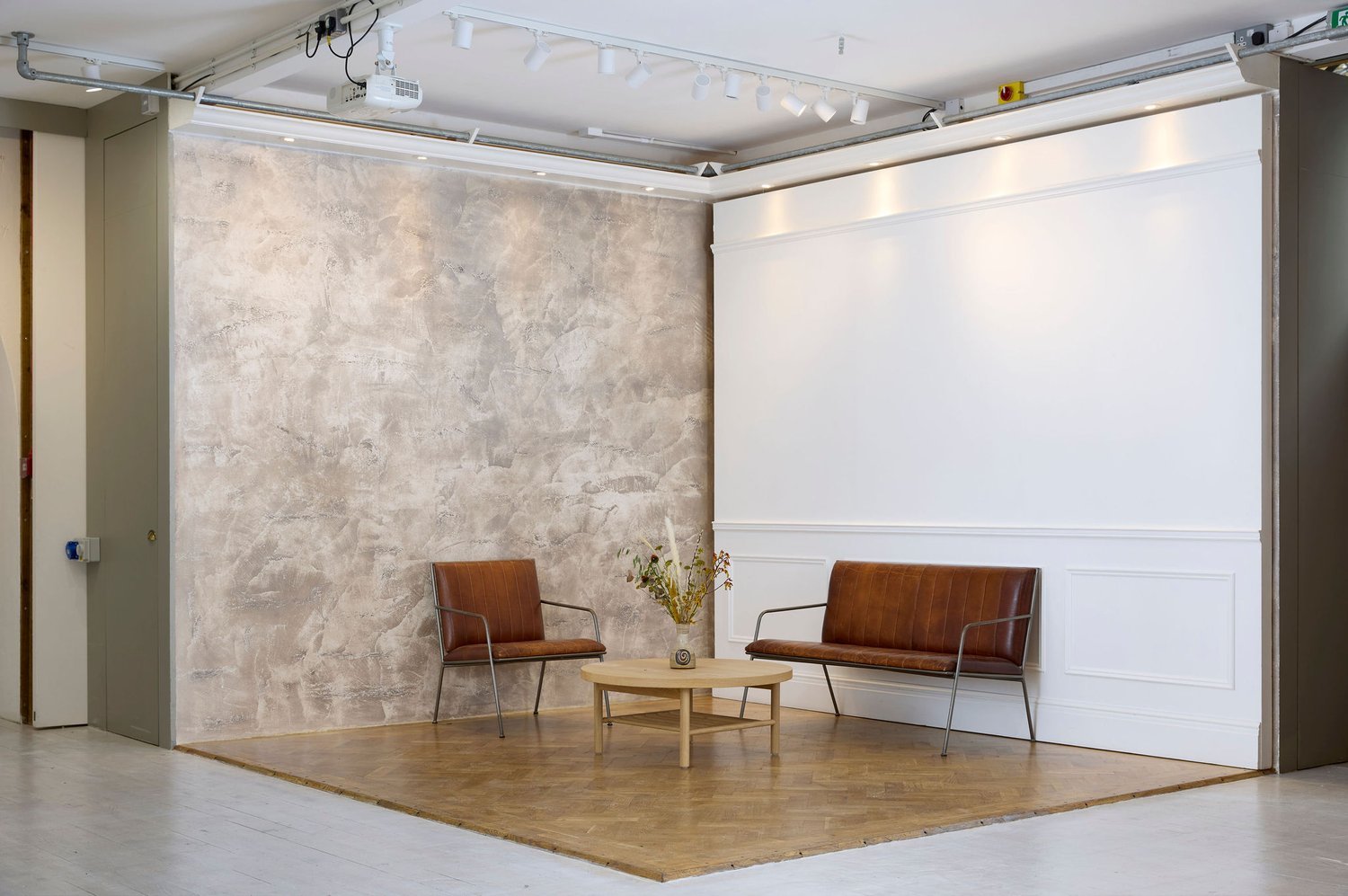 Interior space with two brown leather chairs separated by a small round wooden table with a vase of dried flowers, featuring a textured wall and track lighting in a modern setting.