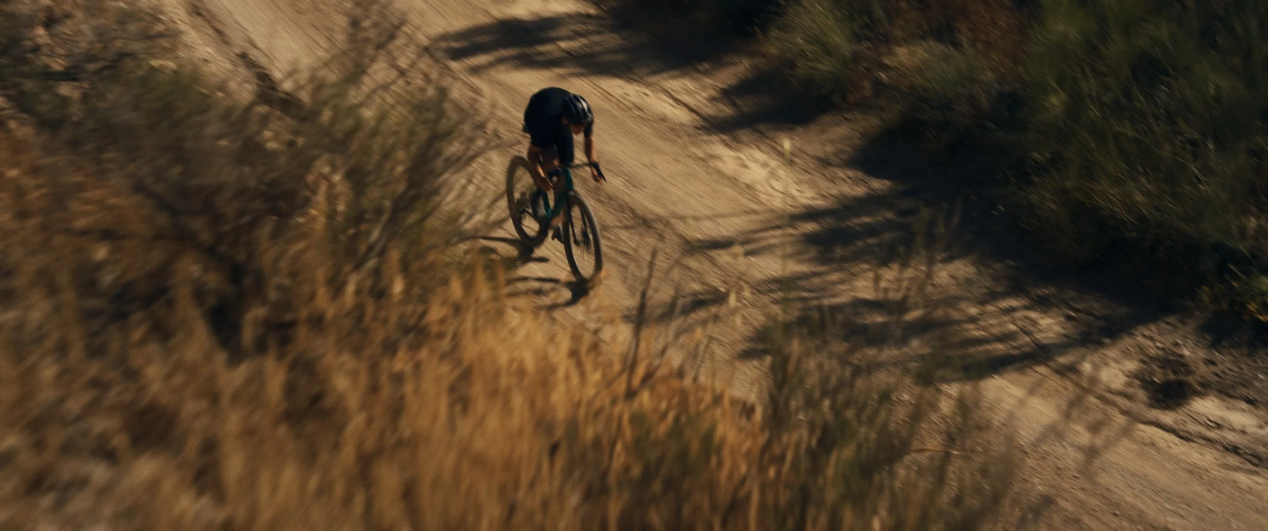Un ciclista in mountain bike che percorre un sentiero sterrato in un ambiente naturale con arbusti e ombre lunghe