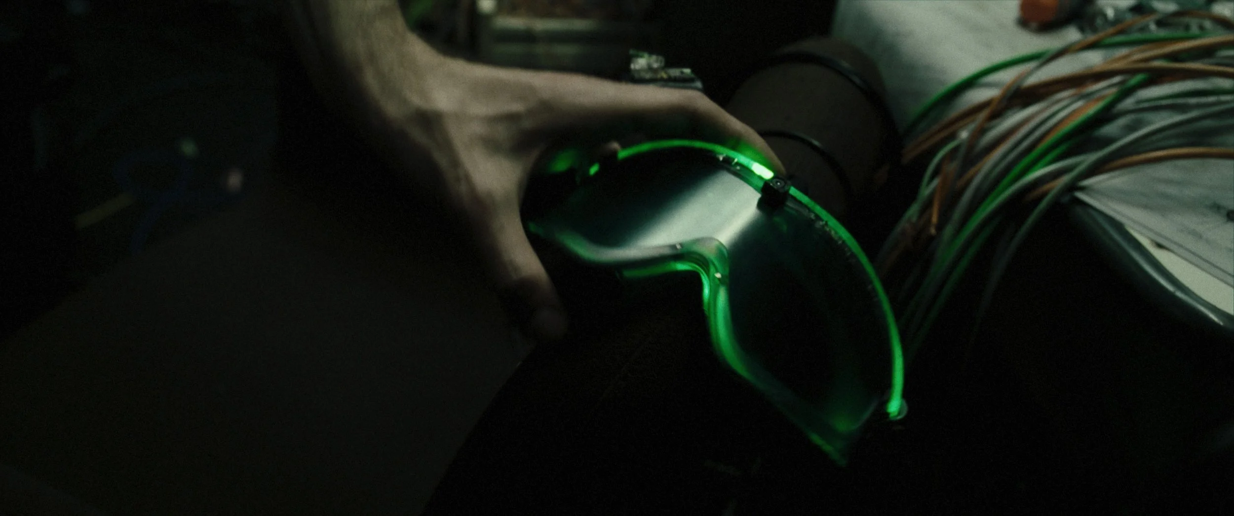 Uno strumento ottico con reticolo illuminato di verde, afferrato da una mano, con cavi e documenti sullo sfondo.