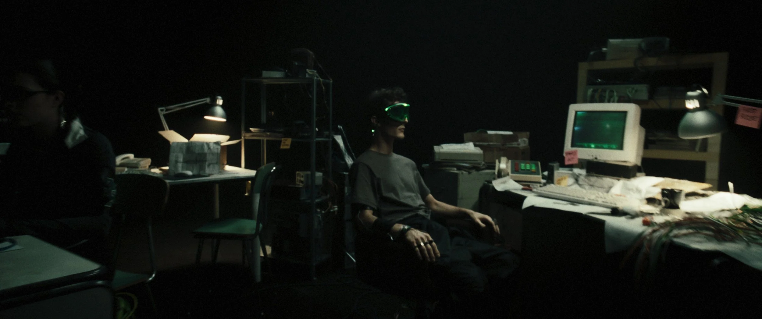 Persona seduta in un ambiente oscuro con attrezzature elettroniche e schermi, indossa occhiali da realtà virtuale o maschera di protezione verde.