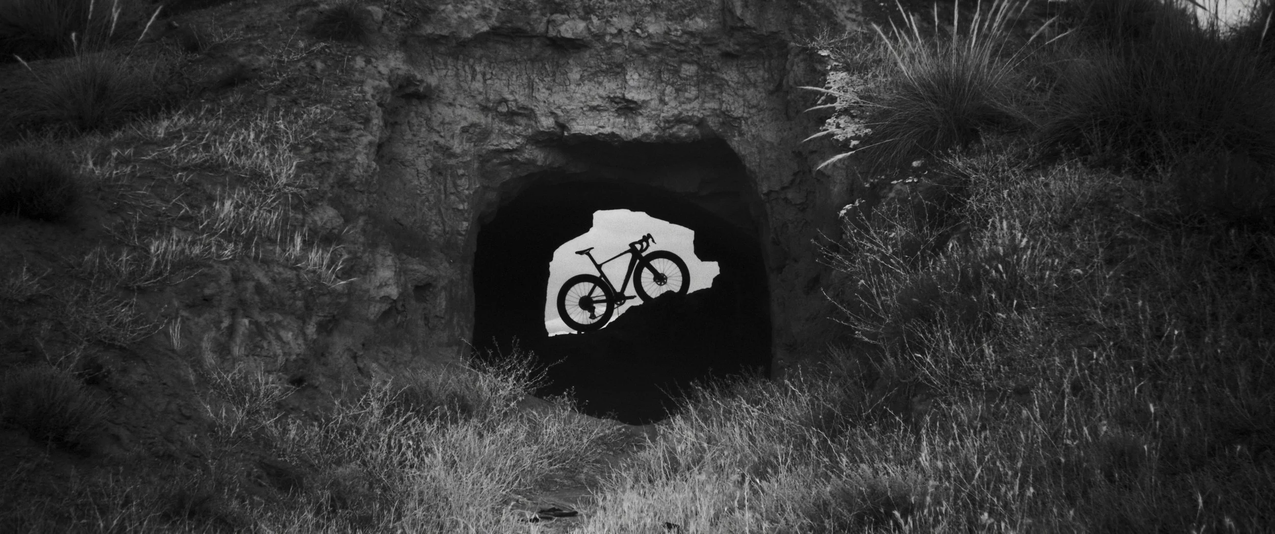 Silhouette di una bicicletta vista attraverso una grotta, circondata da vegetazione. In bianco e nero.