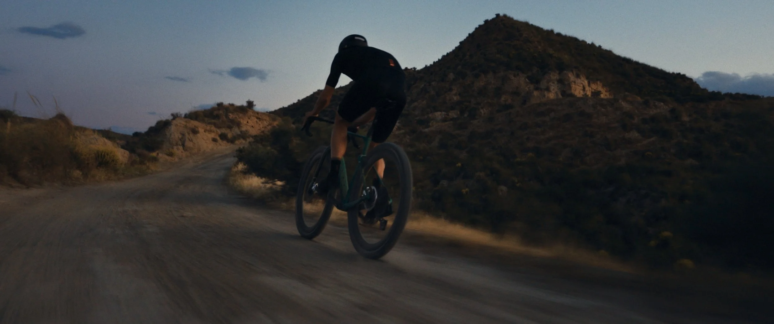 Un ciclista su una strada sterrata in campagna al tramonto, con colline e cielo sereno sullo sfondo.