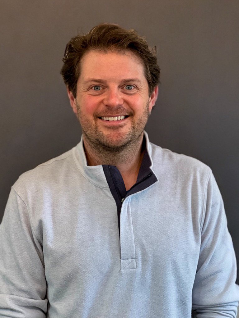 A smiling man with brown hair and blue eyes, wearing a light gray quarter-zip pullover, standing against a plain dark background.