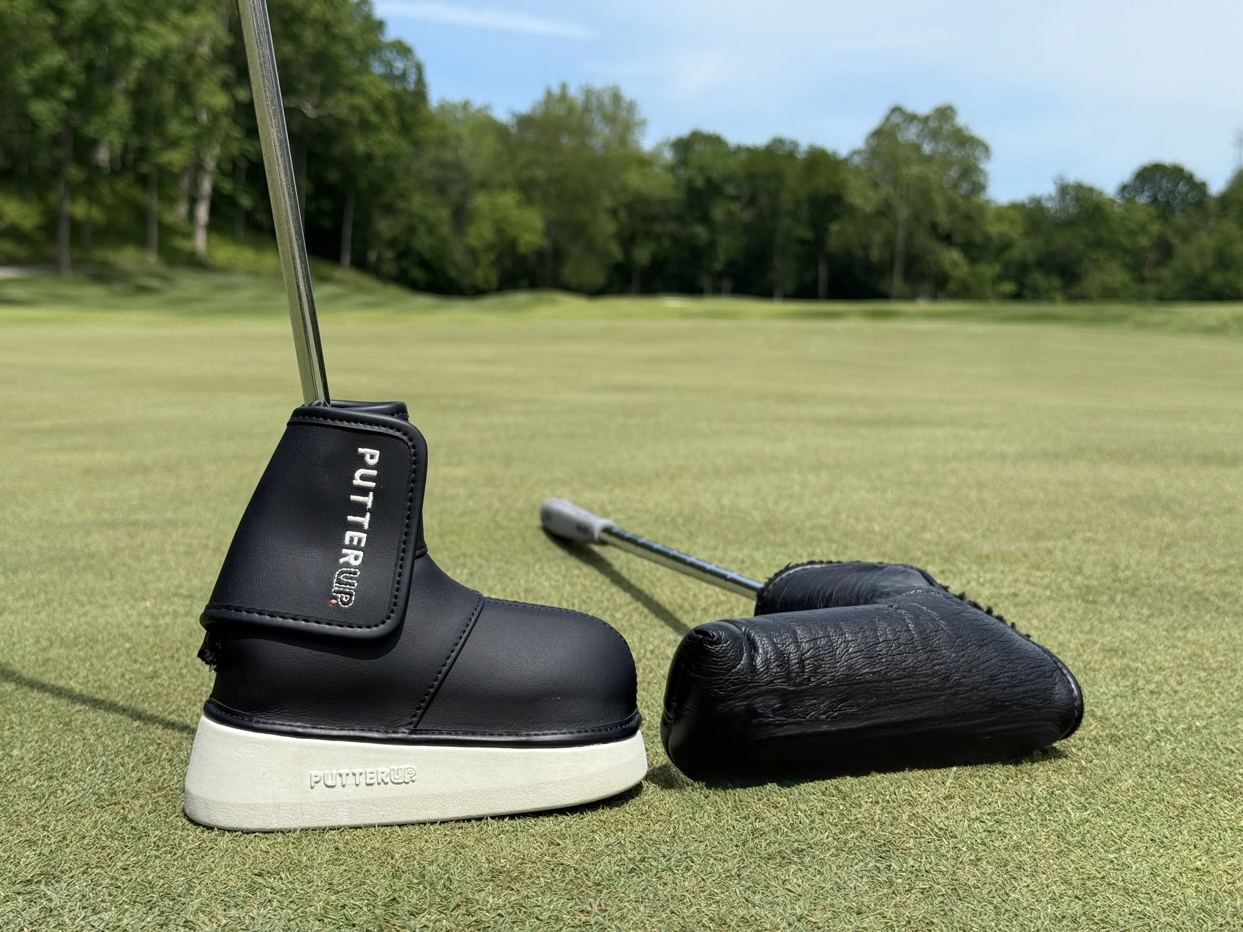 Golf putter with protective cover on a golf course green, compared to regular putter cover on the ground, with trees and blue sky in the background.