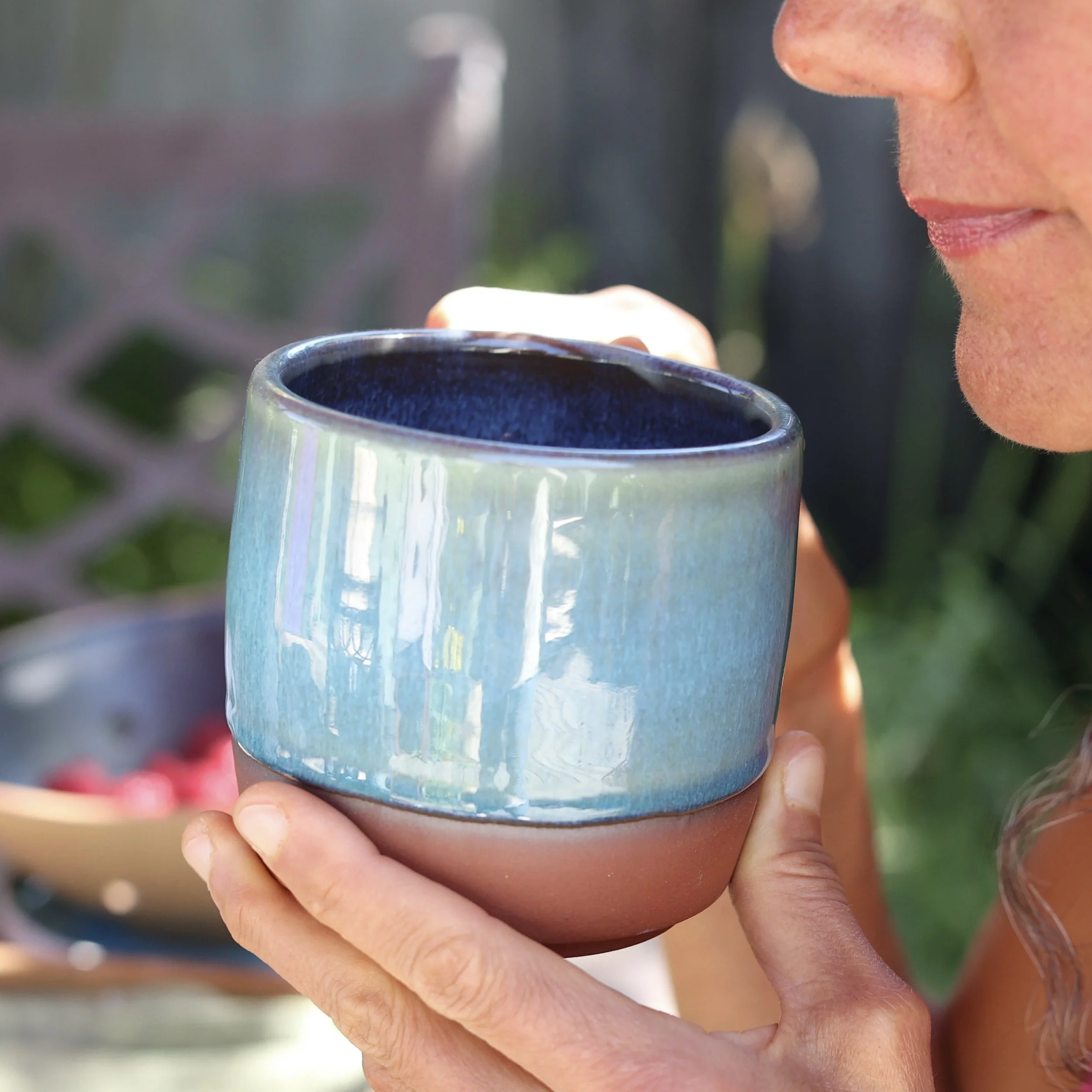 A woman holding a ceramic bowl, possibly filled with a beverage, with a blurred outdoor background.