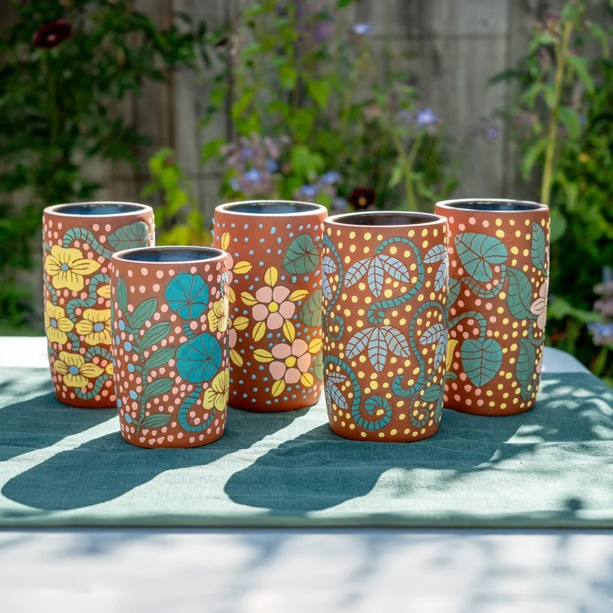 Six colorful hand-painted ceramic cups with floral and vine patterns arranged outdoors on a table with a green tablecloth, casting shadows in sunlight.