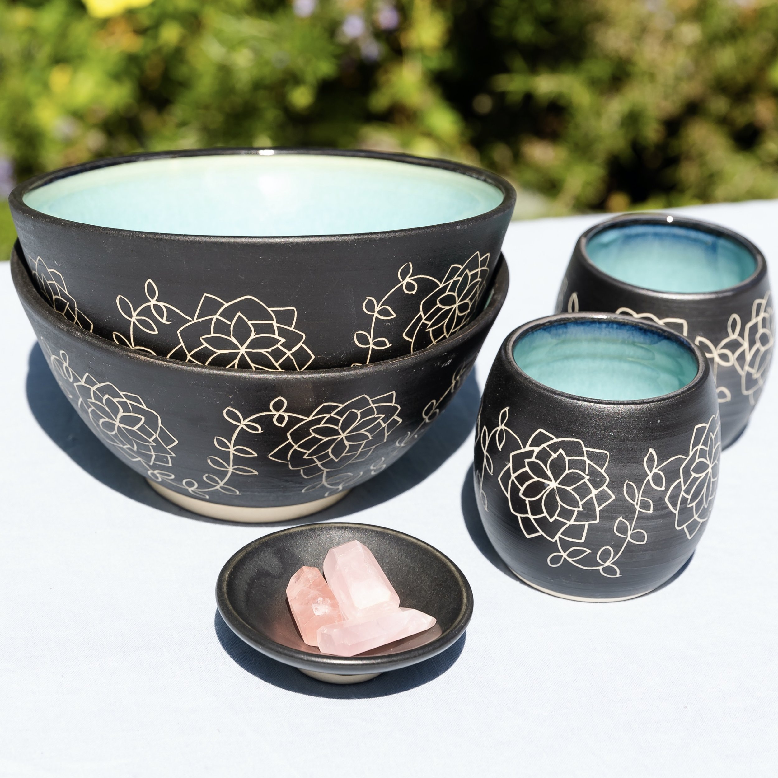 Set of handcrafted ceramic bowls and cups with floral designs, displayed on a table outdoors with sunlight and prayer flags in the background.