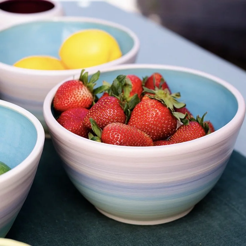Set of colorful, glazed ceramic bowls and a tall vase with pastel striped pattern on a dark cloth outdoors with greenery in the background.