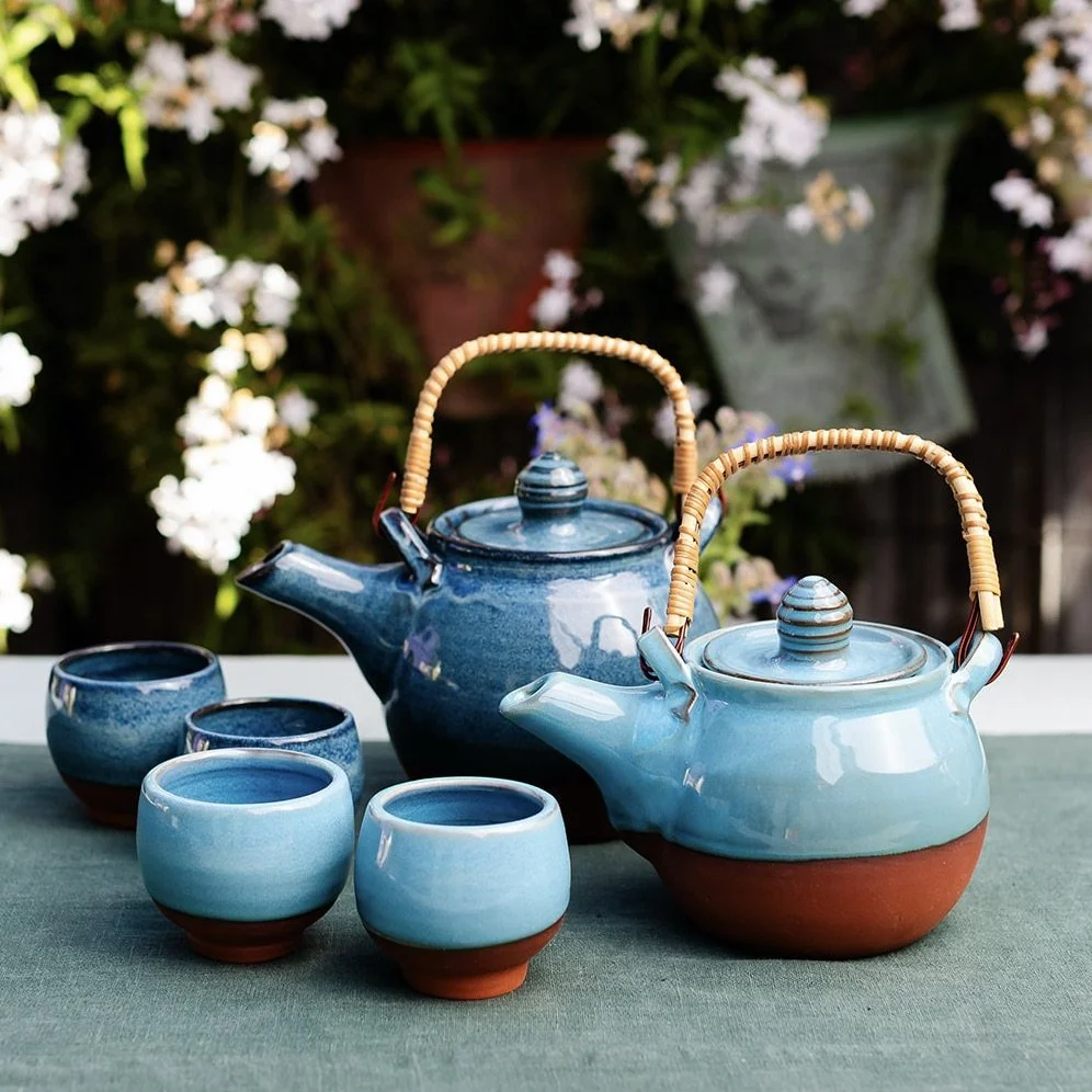Black ceramic teapot with a rattan handle and two matching cups on a white table, with a blue and white blurred water background.