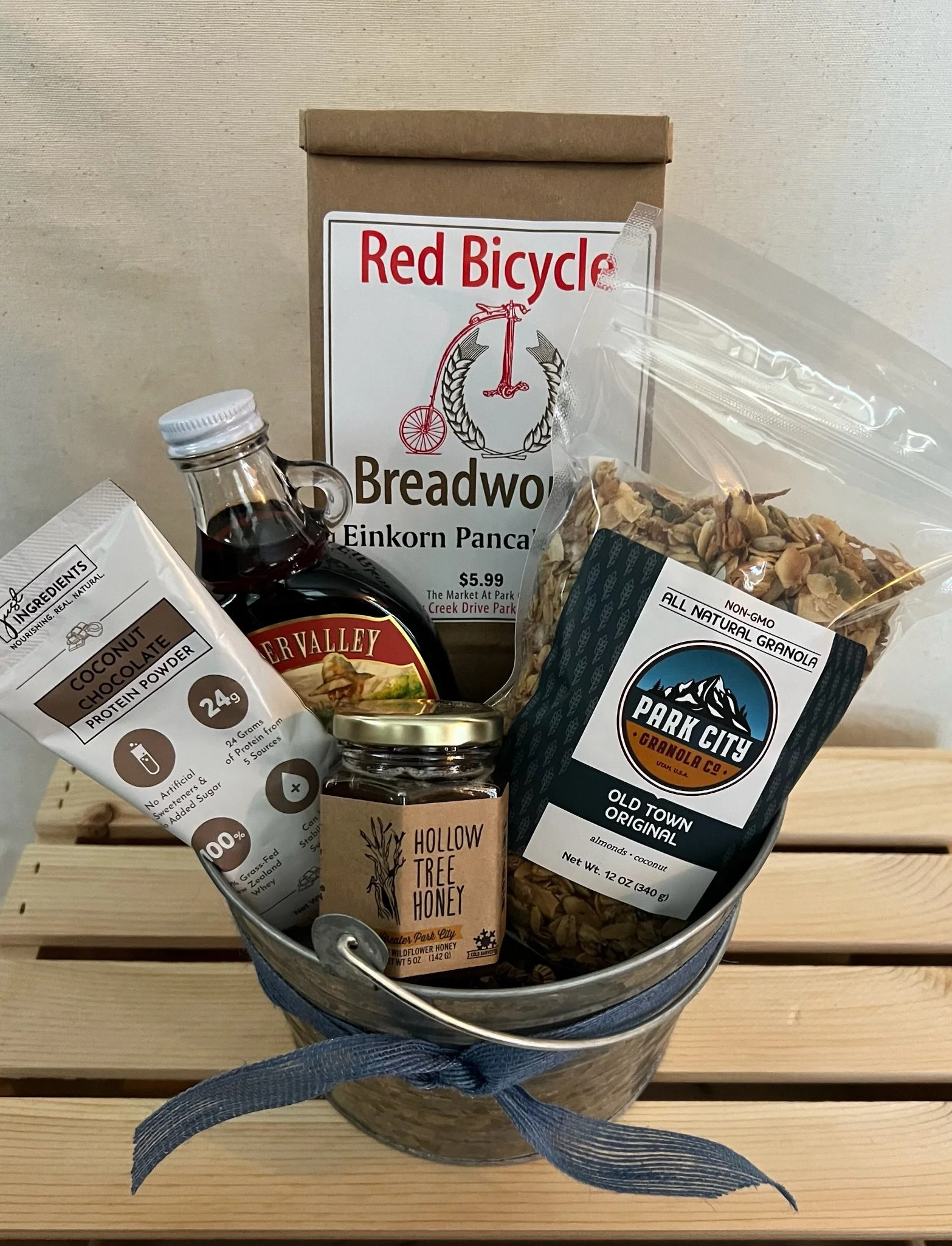 A metal bucket containing various food items including a box of Red Bicycle bread, a bag of Park City granola, a bottle of maple syrup, and a box of Hollow Tree Honey, all set on a wooden surface.