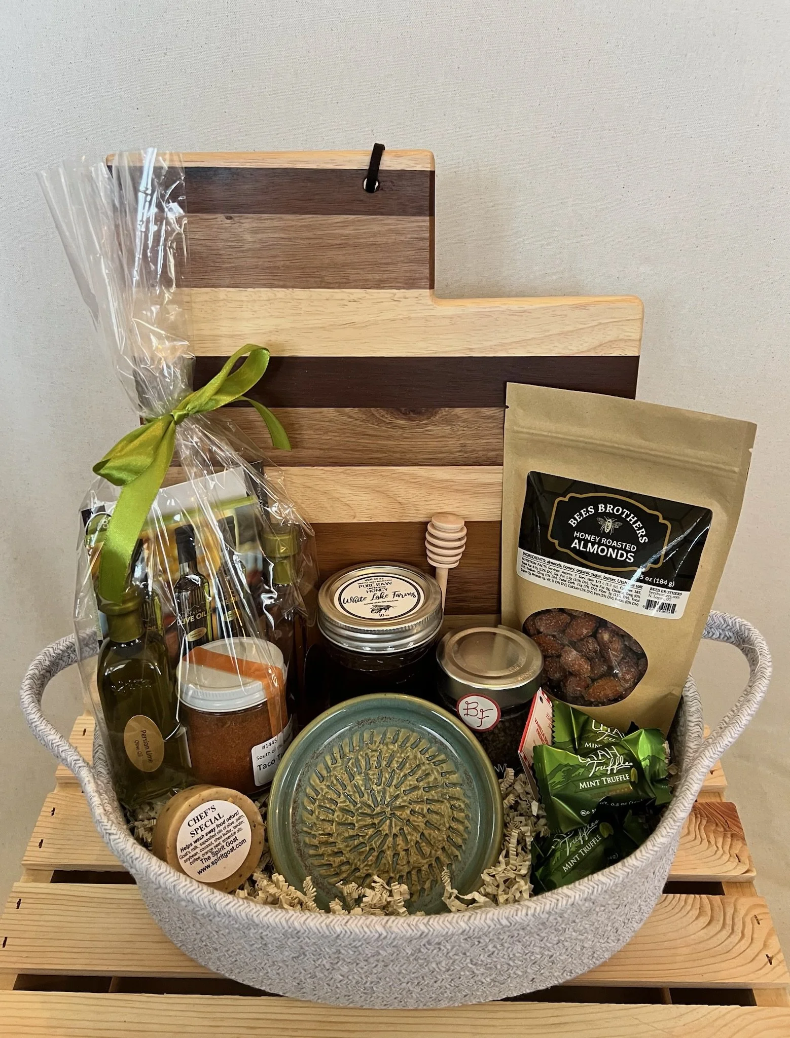 A gift basket containing various food items including a sealed bag of honey roasted almonds, a jar of honey, a bag of mint truffle chocolates, and some bottles of olive oil. The basket is decorated with wood and paper elements and placed on a wooden surface.