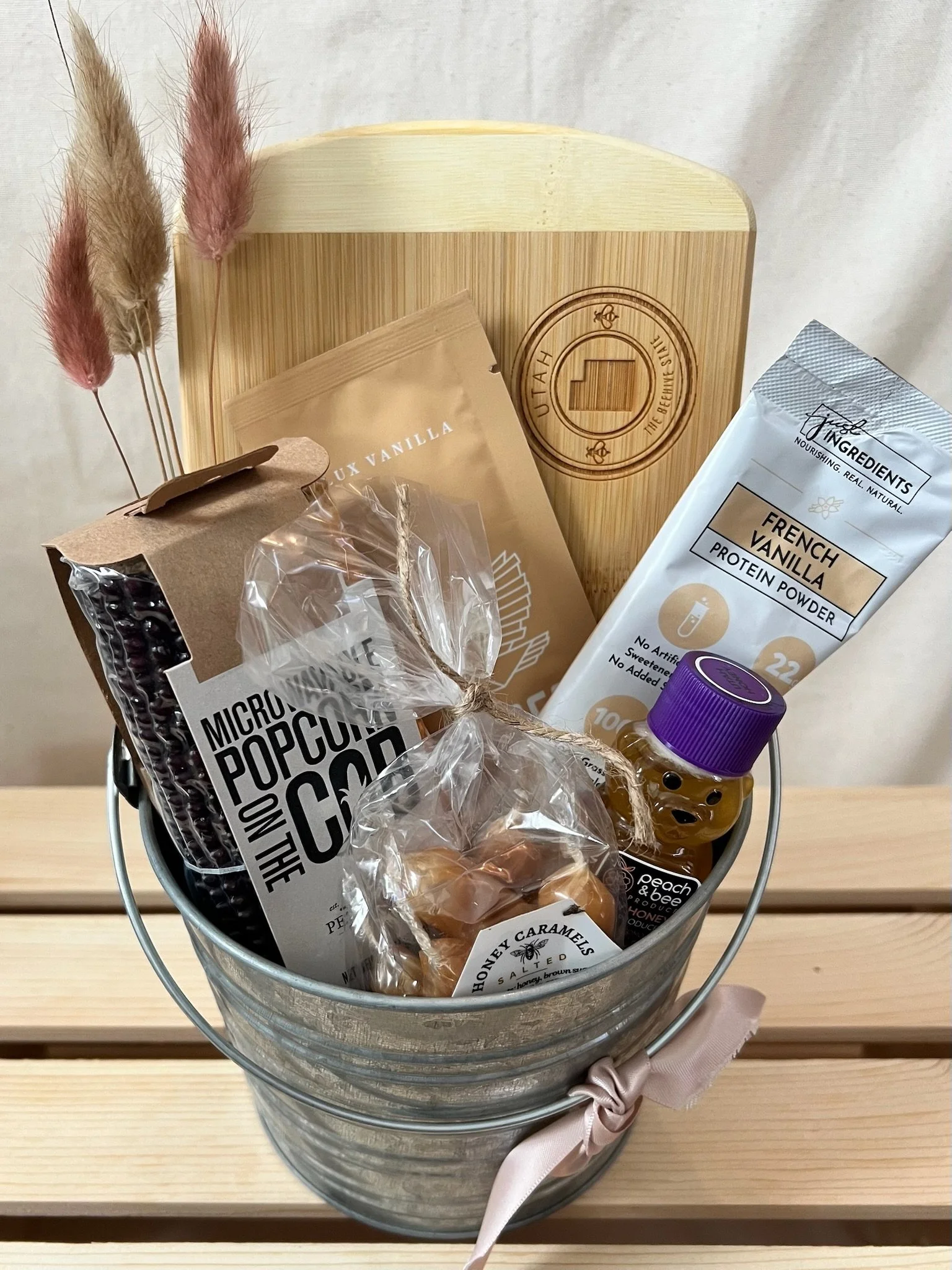 A metal gift basket containing various food items and treats, decorated with a pink ribbon, sitting on a wooden surface.