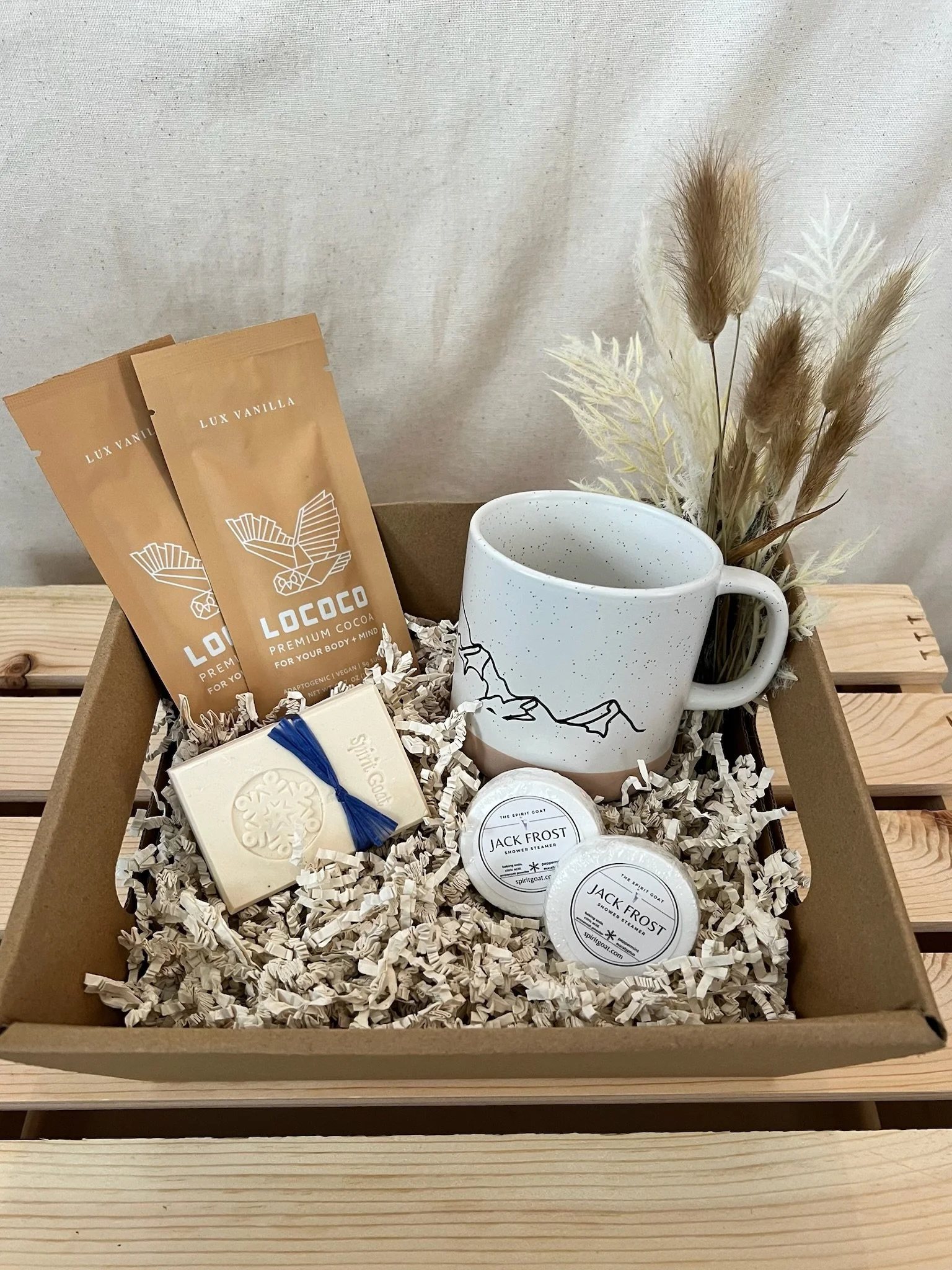 Gift box with a mug, packets of premium cocoa, soap, and small containers of shower steamer, with dried decorative plants inside a box filled with shredded paper, on a wooden surface.