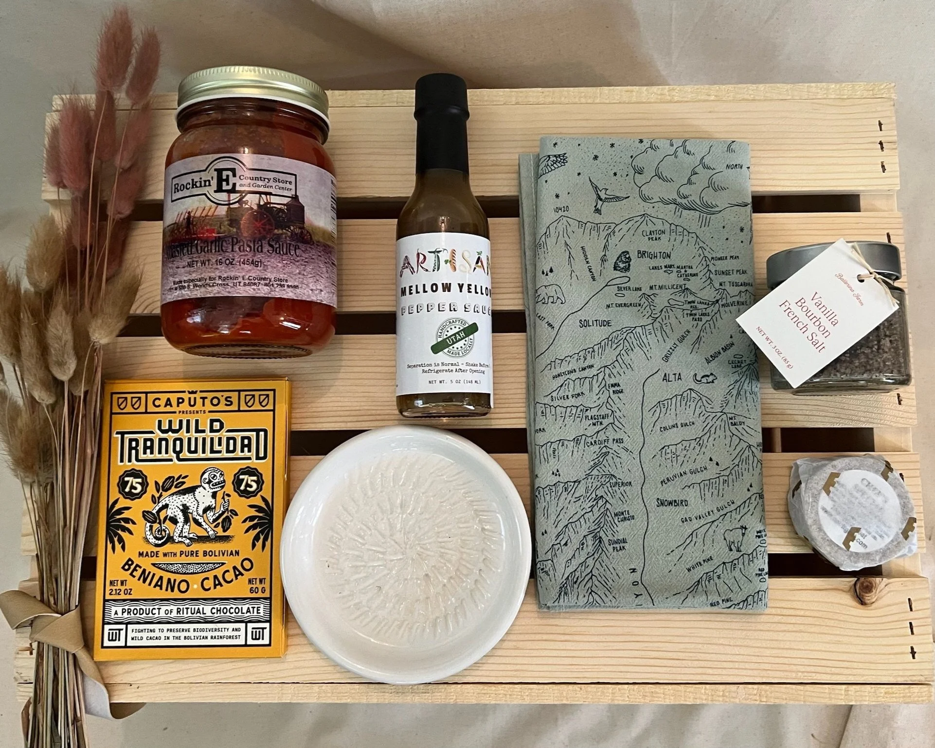A wooden tray holding a jar of roasted garlic pasta sauce, a bottle of mellow yellow peppered sauce, a blue map with mountain names, a small jar of vanilla bourbon French salt, a yellow packet of Beniano cacao, a white ceramic plate, and some dried plants on the left side.