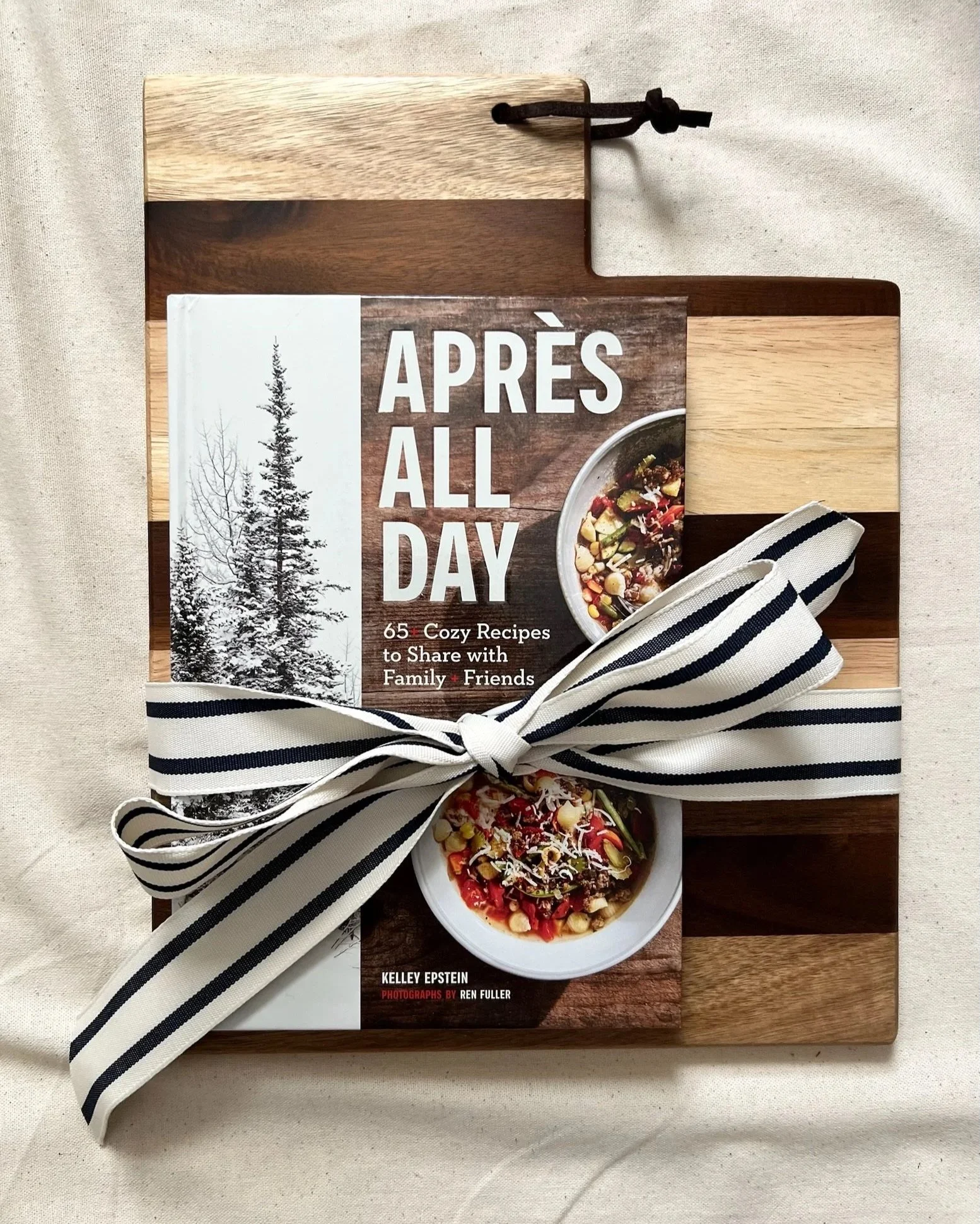 A book titled "Après All Day" resting on a striped black and white ribbon, placed on a wooden cutting board with a small bowl of salad nearby. The background is a fabric surface.