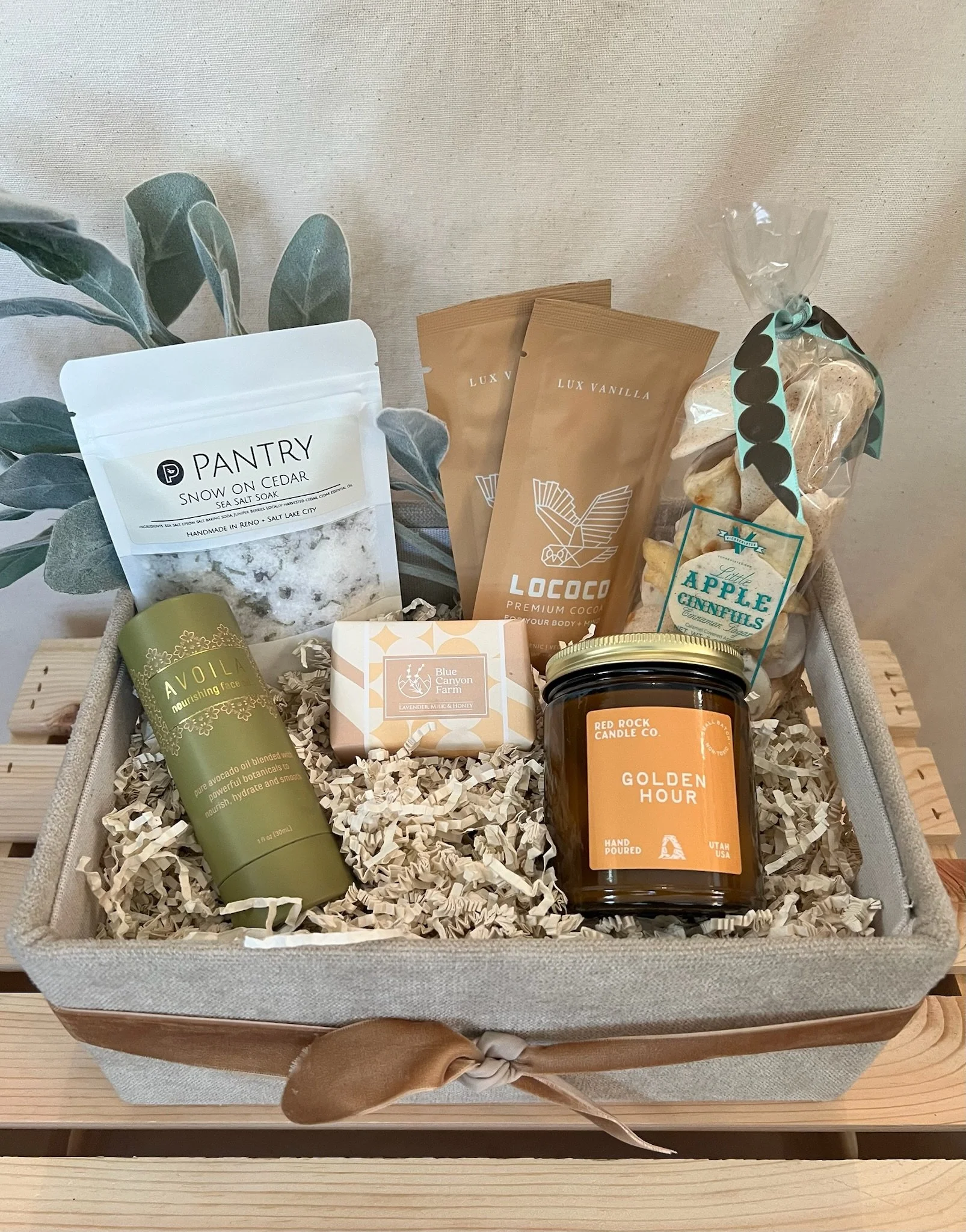 A gift basket containing skincare products, chocolate, candles, and snacks on a wooden surface with a beige fabric background.