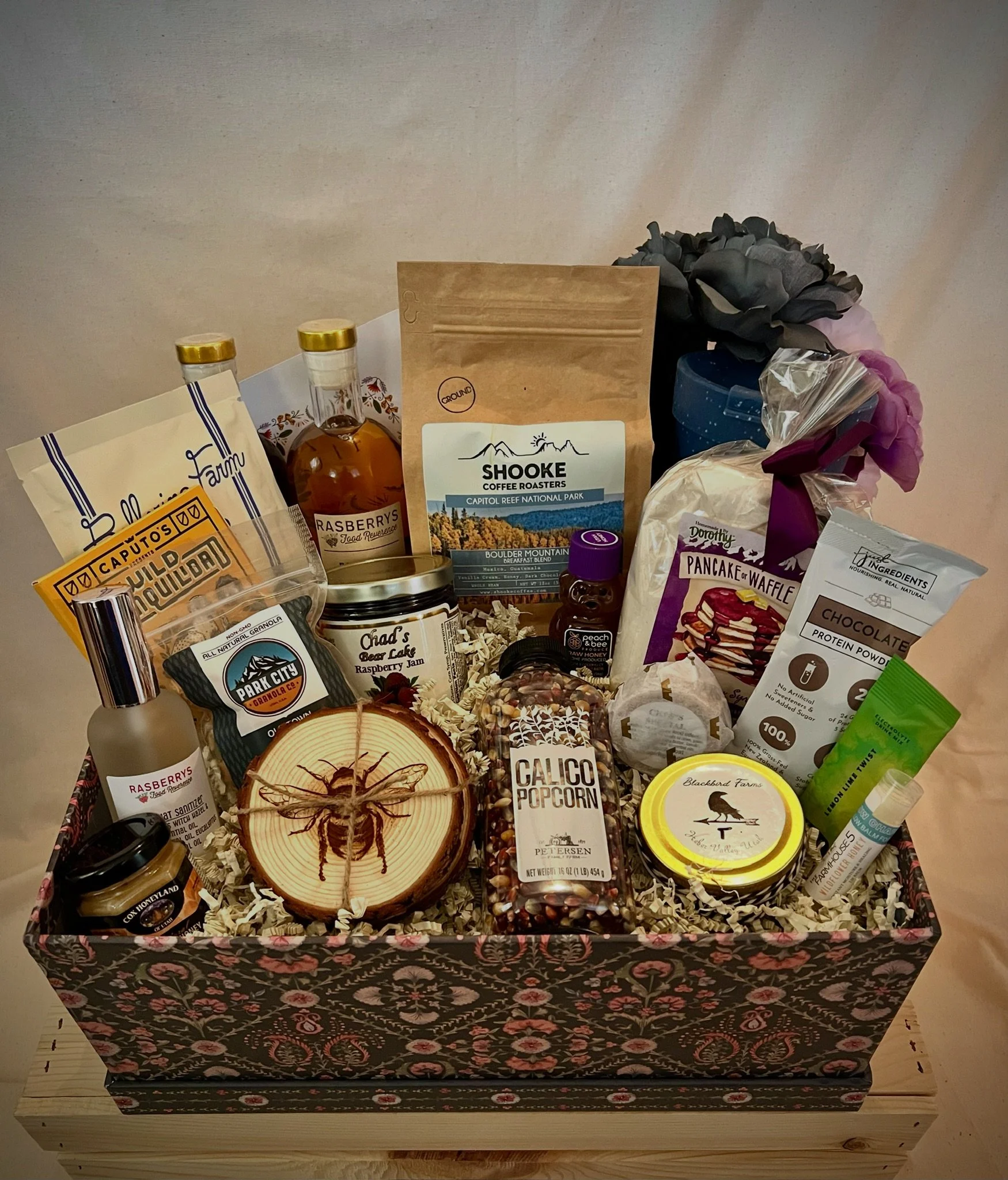 A gift basket containing various food and beverage items, including jars of honey, packets of coffee, snacks, and a bouquet of flowers, all arranged on a wooden surface.