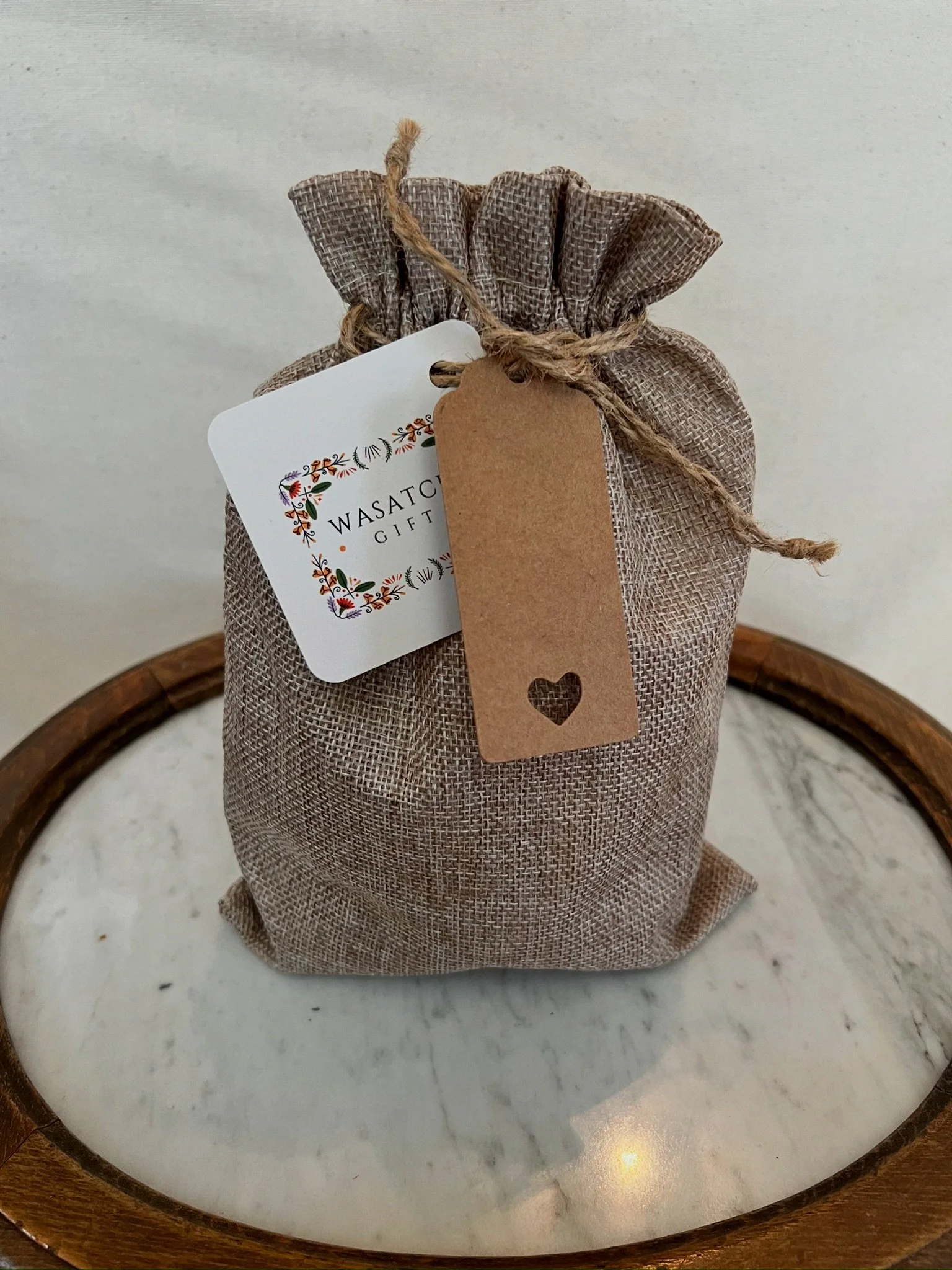 A linen drawstring bag with a tag and a gift card inside, placed on a round wooden tray. The tag has a heart cut-out, and the gift card features decorative floral artwork and the words 'WASATCH GIFT'.