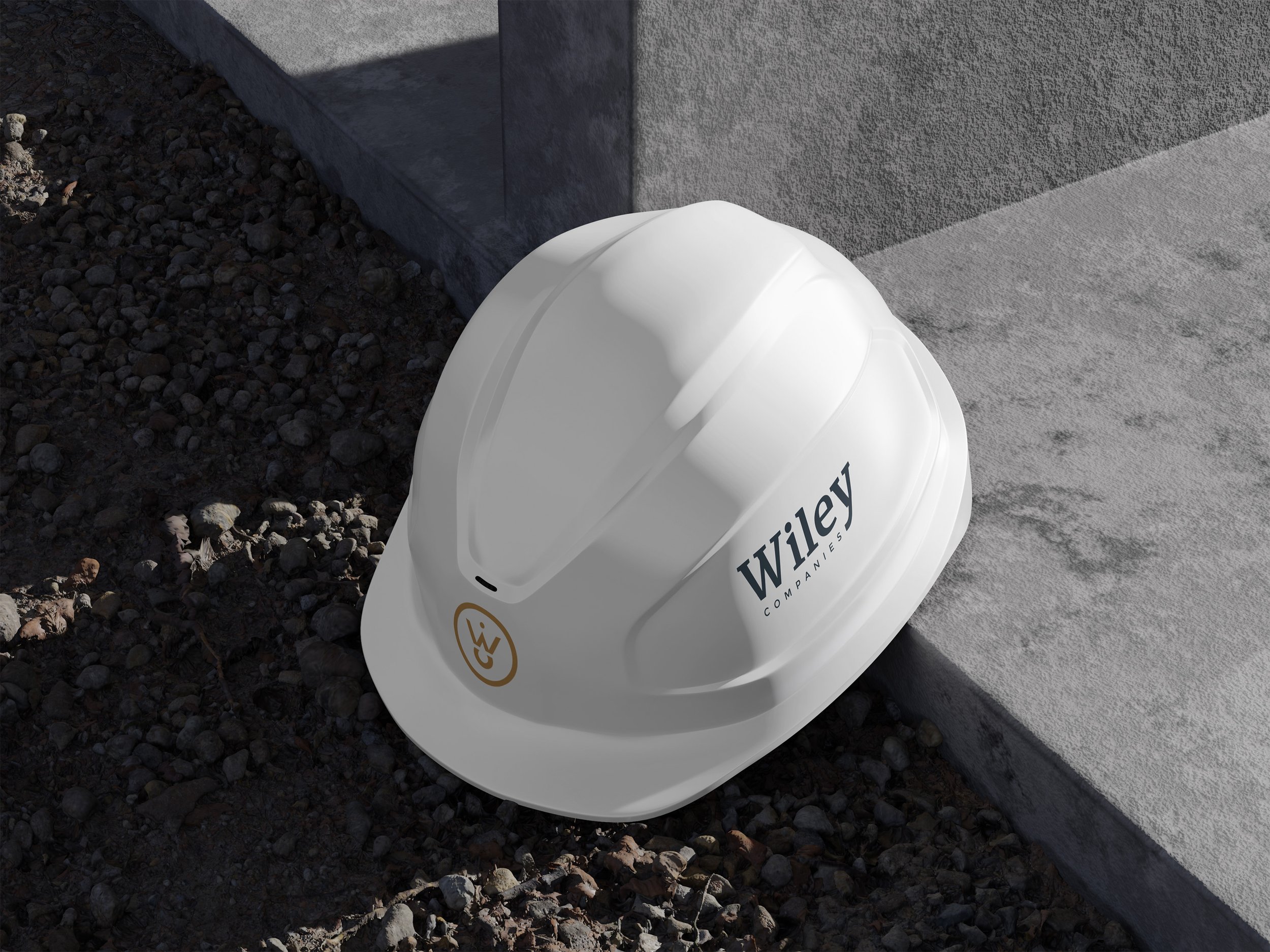 White construction helmet with the Wigley Company logo in black, resting on rocky ground next to a concrete step.