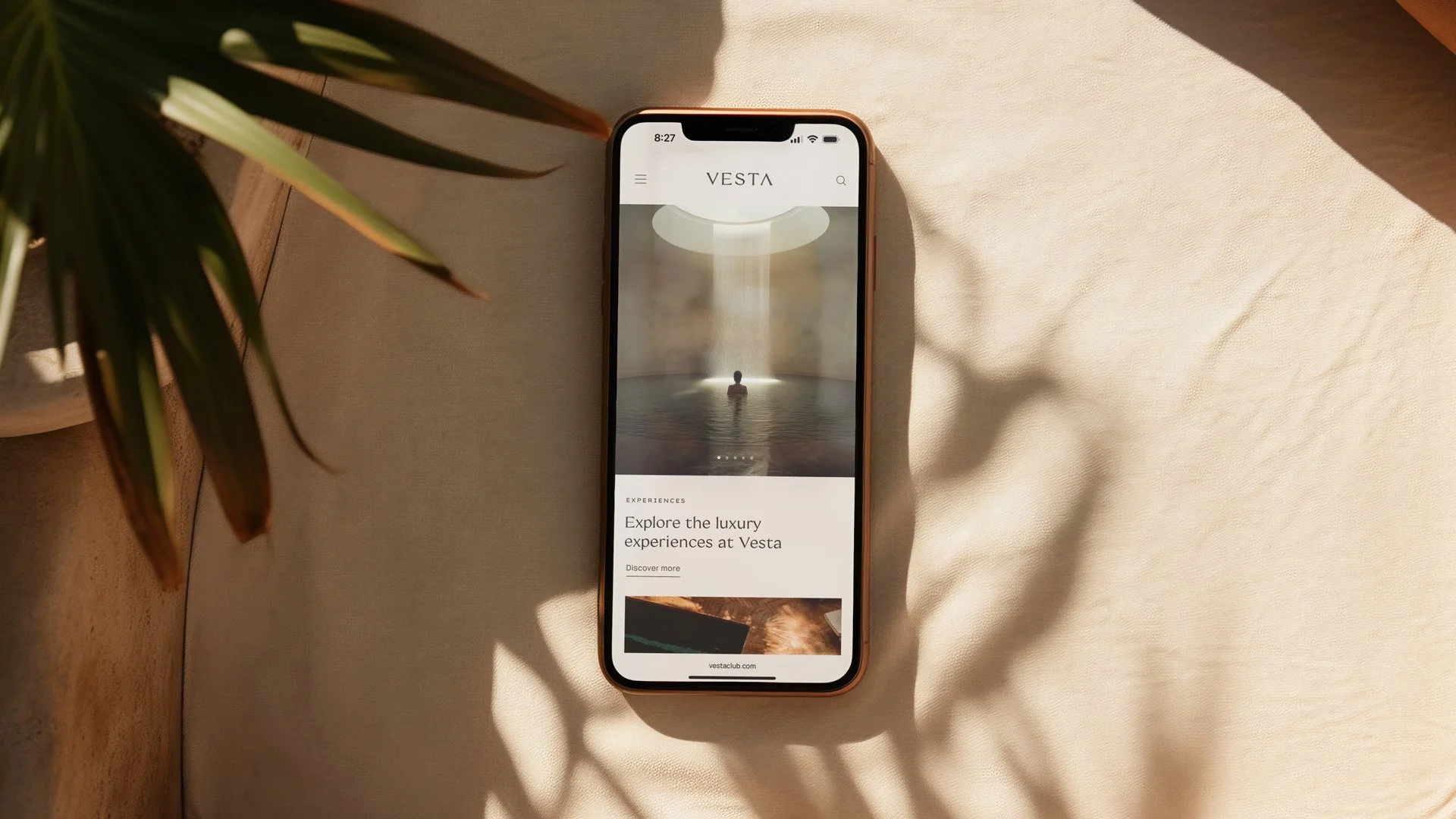 Smartphone on a light-colored cloth surface displaying a website called Vesta with an image of a person in a fountain with a skylight above, with shadows of plant leaves on the cloth.