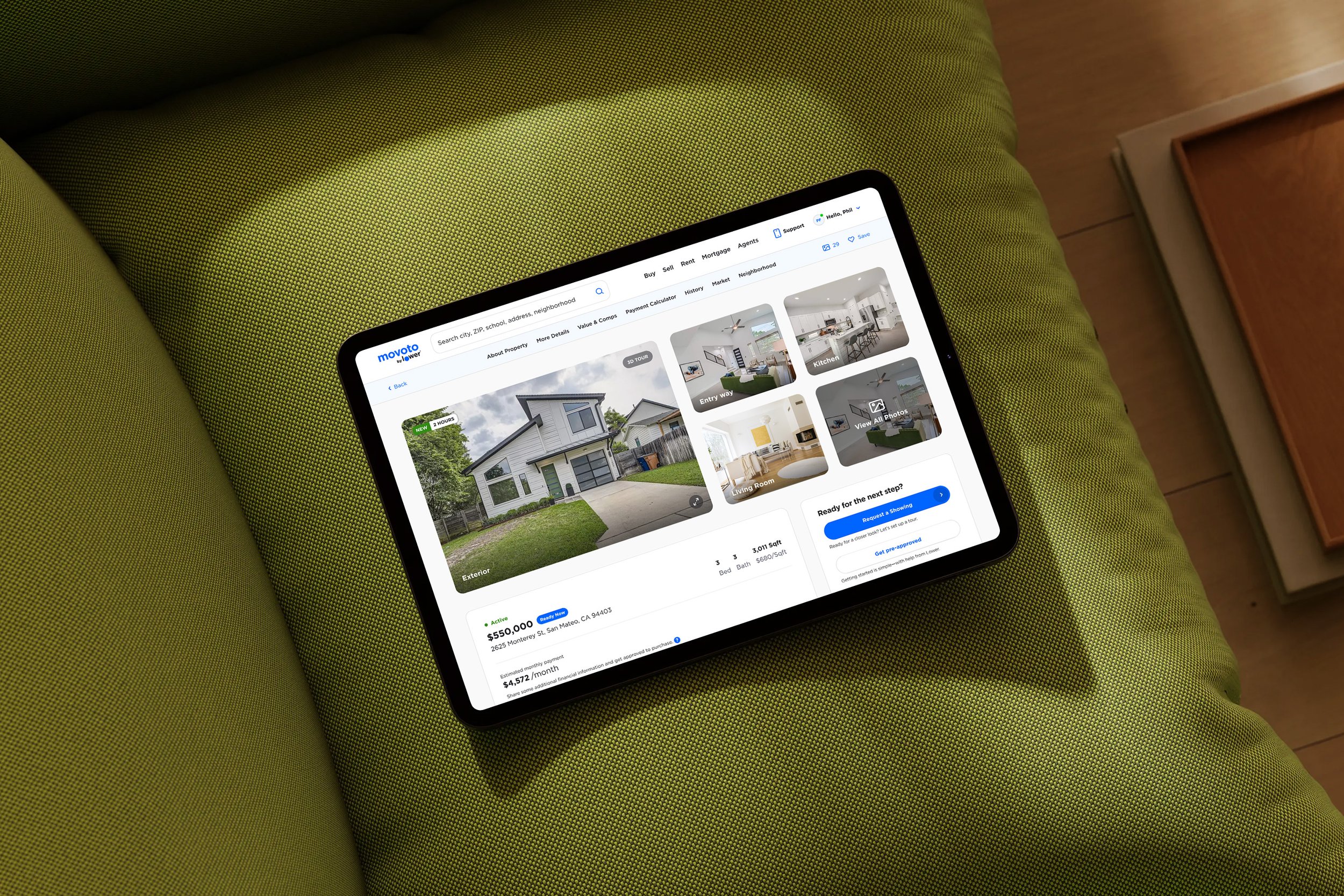 iPad displaying a real estate listing for a modern white house with a driveway, placed on a green upholstered surface with a wooden floor nearby.