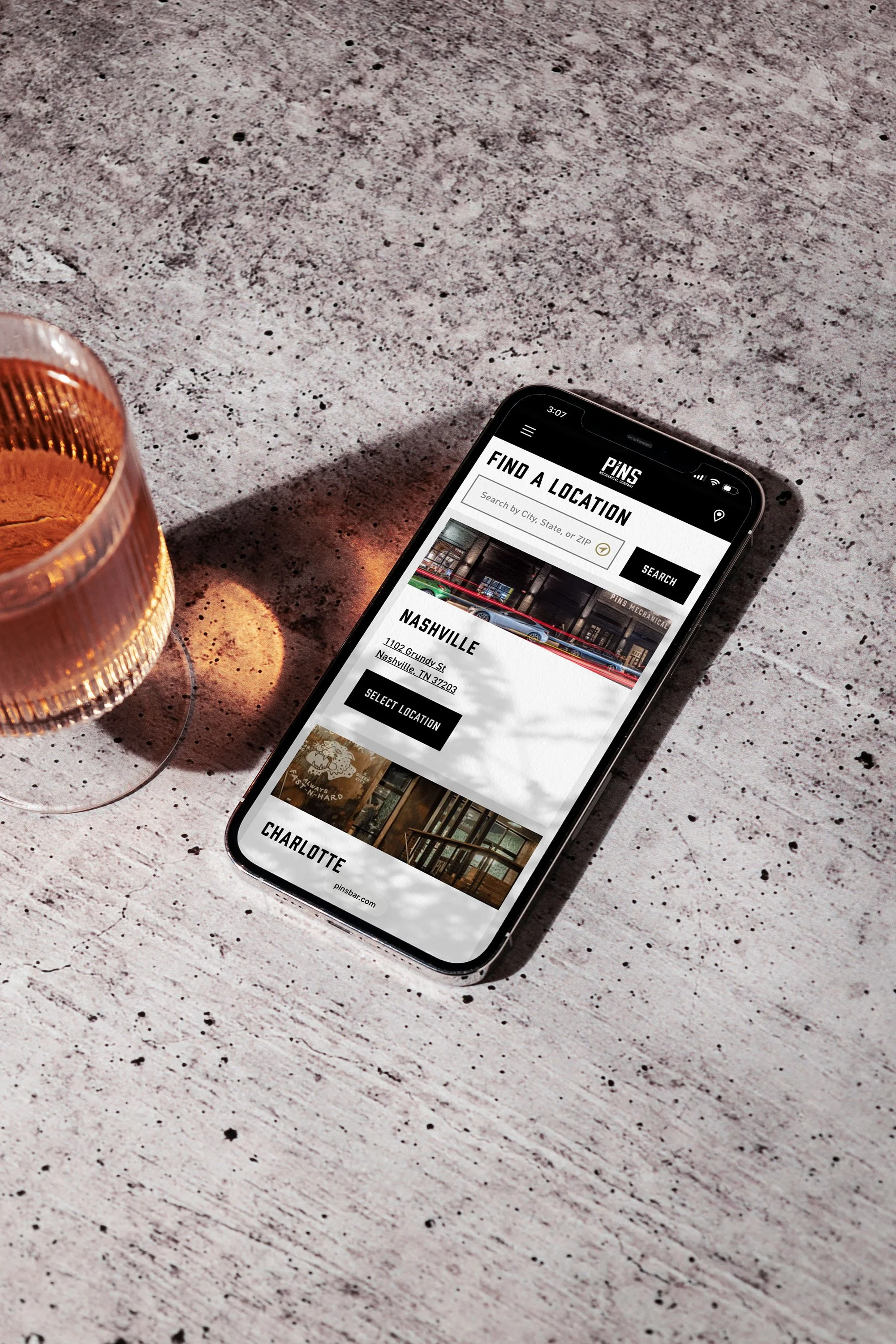 Smartphone on a table displaying a Pins app with location options for Nashville and Charlotte, and a glass of amber-colored beverage nearby.