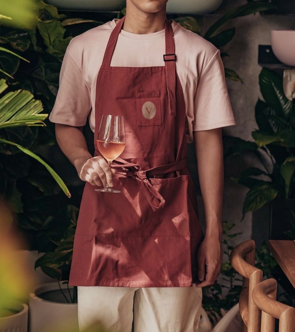 A person wearing a light-colored T-shirt and a red apron holding a glass of rosé wine in a setting with green plants.