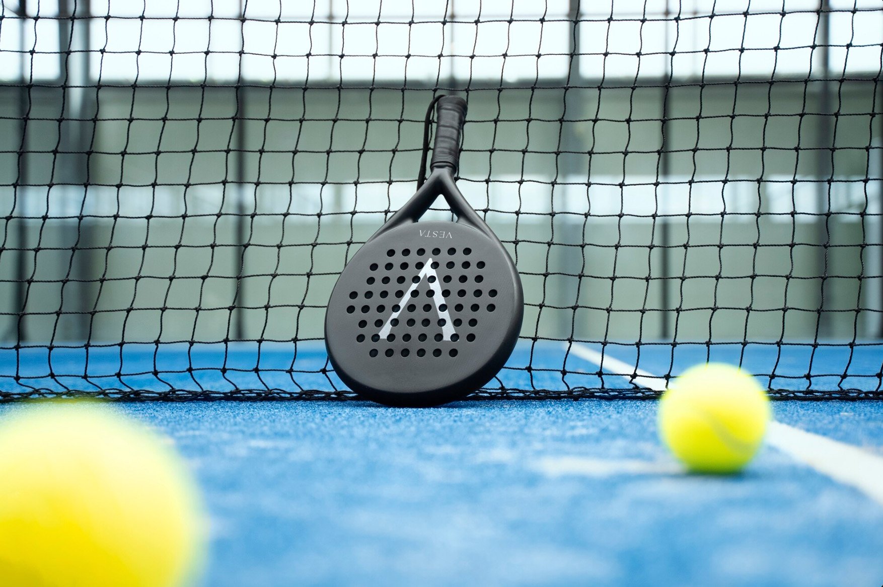 Tennis racket leaning against a tennis net on a blue court with two yellow tennis balls in the foreground.