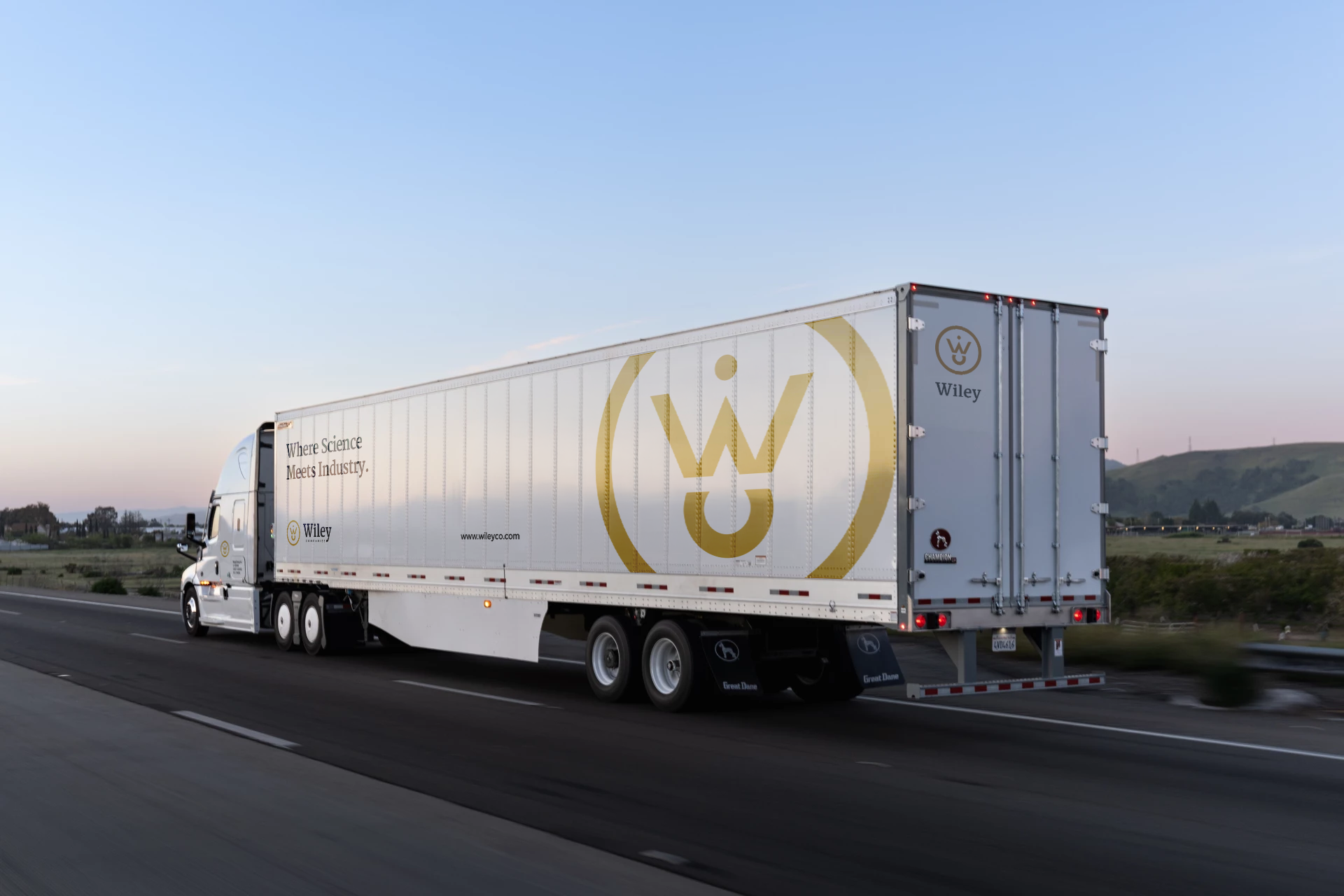 A white semi-truck with Wiley branding, driving on a highway during sunset, with a scenic rural landscape in the background.