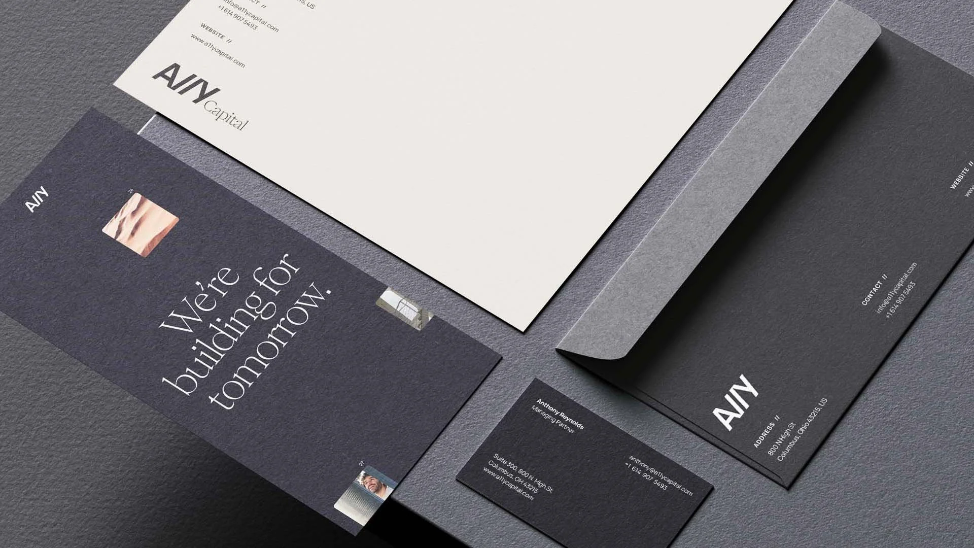 Layout of corporate stationery, including a dark-colored business card, a white sheet of paper, and a black envelope with white text, all on a gray textured surface.