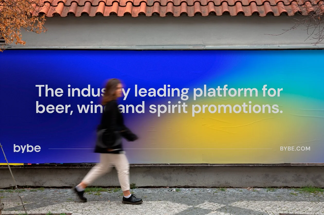 A woman walking past a large colorful billboard on a city sidewalk. The billboard promotes a platform for beer, wine, and spirit promotions, with the text 'The industry leading platform for beer, wine and spirit promotions.' The background of the billboard features a gradient of blue, teal, and yellow colors, and the words 'bybe' and 'BYBE.COM' are visible on it.