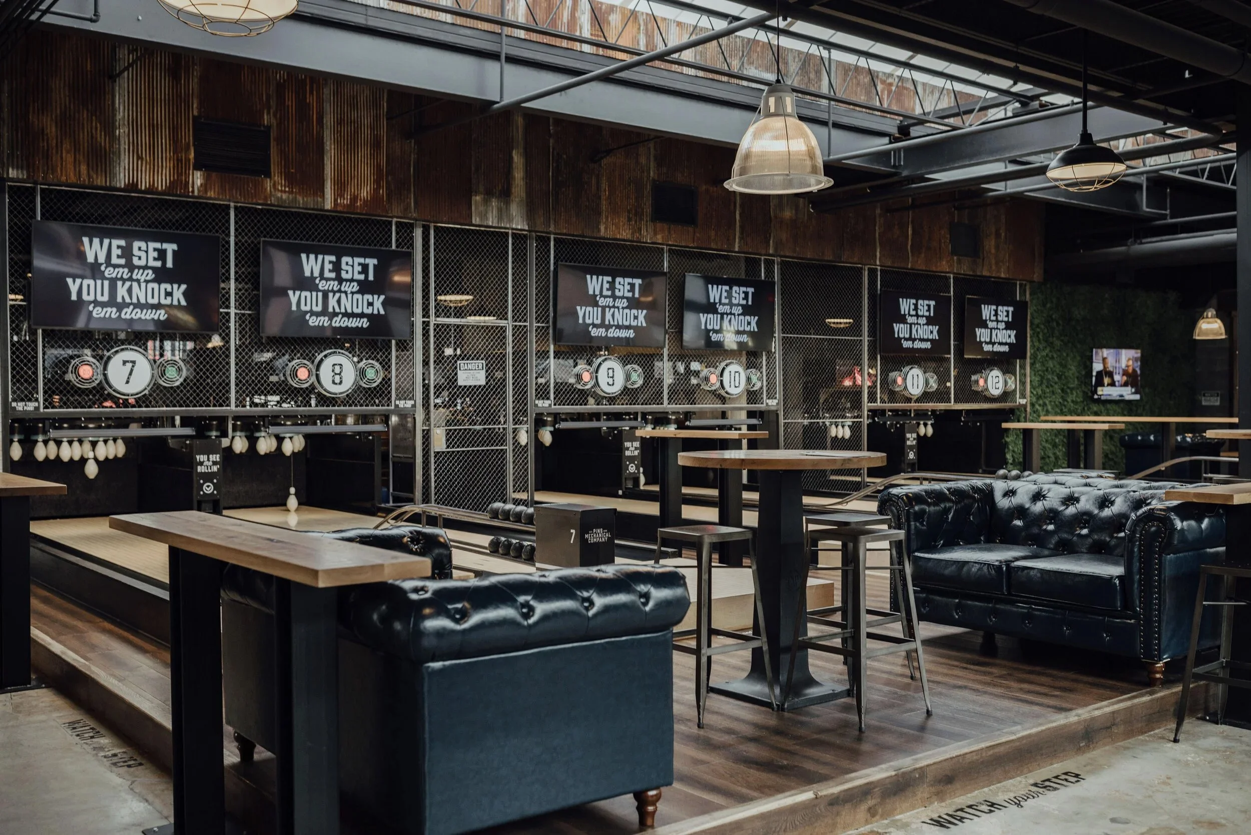 Interior of a modern bowling alley with black leather sofas, high tables, and digital score screens displaying 'We set 'em up, you knock 'em down'.