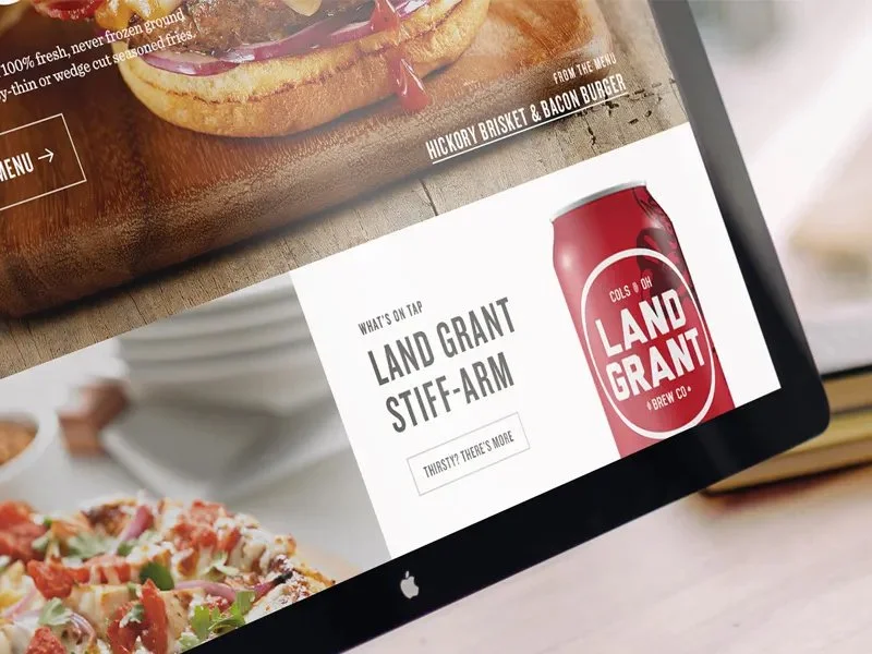 Computer screen displaying a promotional webpage for Land Grant Stif-Arm, a beverage from Land Grant Brew Co., with a can of the drink depicted. The page also features food images including a burger and a pizza.