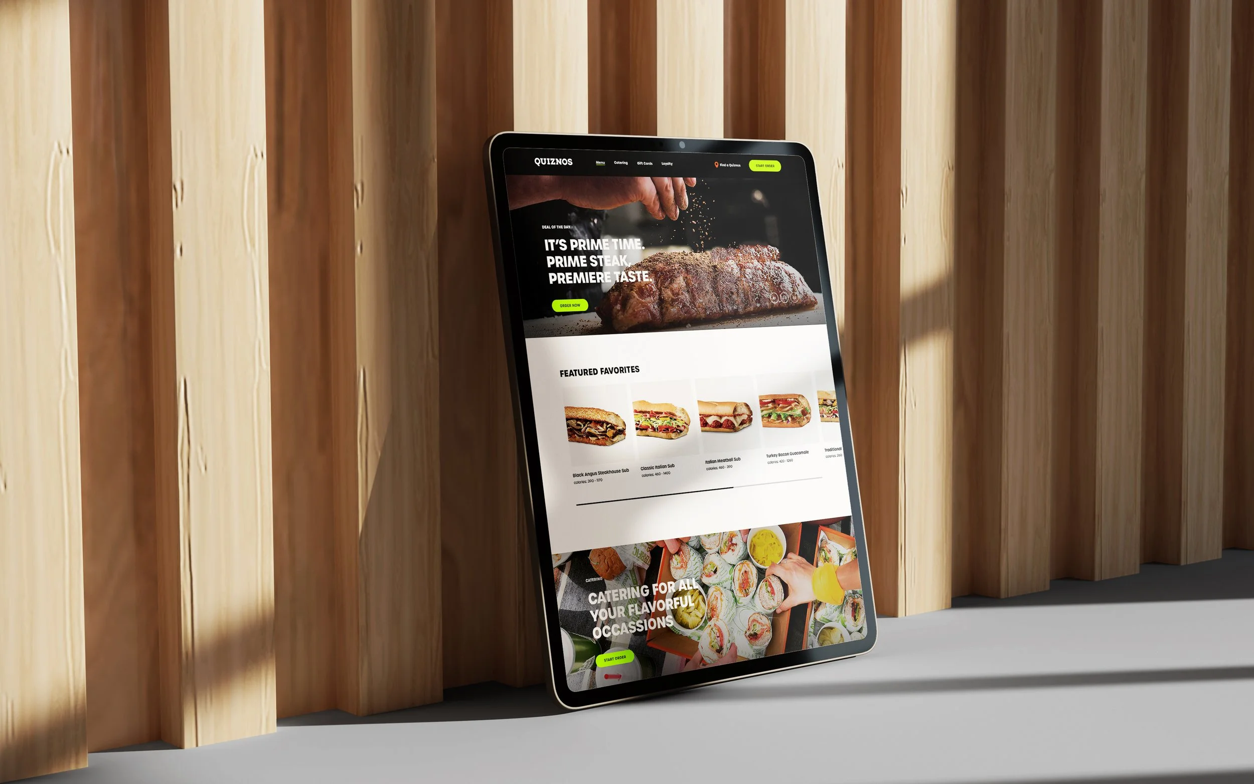 A tablet leaning against a wooden wall displaying a food website with images of sandwiches, sushi, and a steak.