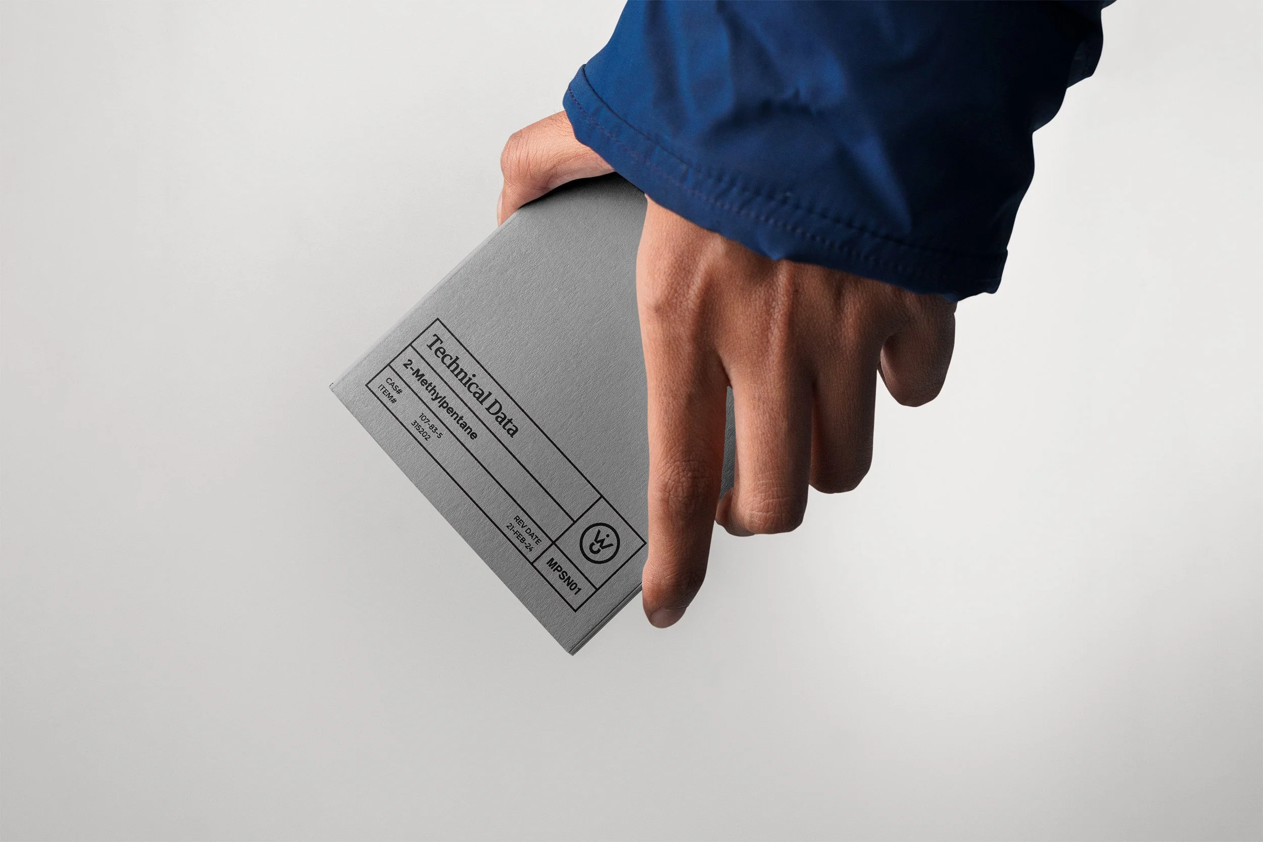 A person wearing a blue jacket holding a rectangular gray box labeled 'Technical Data' with chemical information for '2-Methoxyphenetole' against a plain gray background.