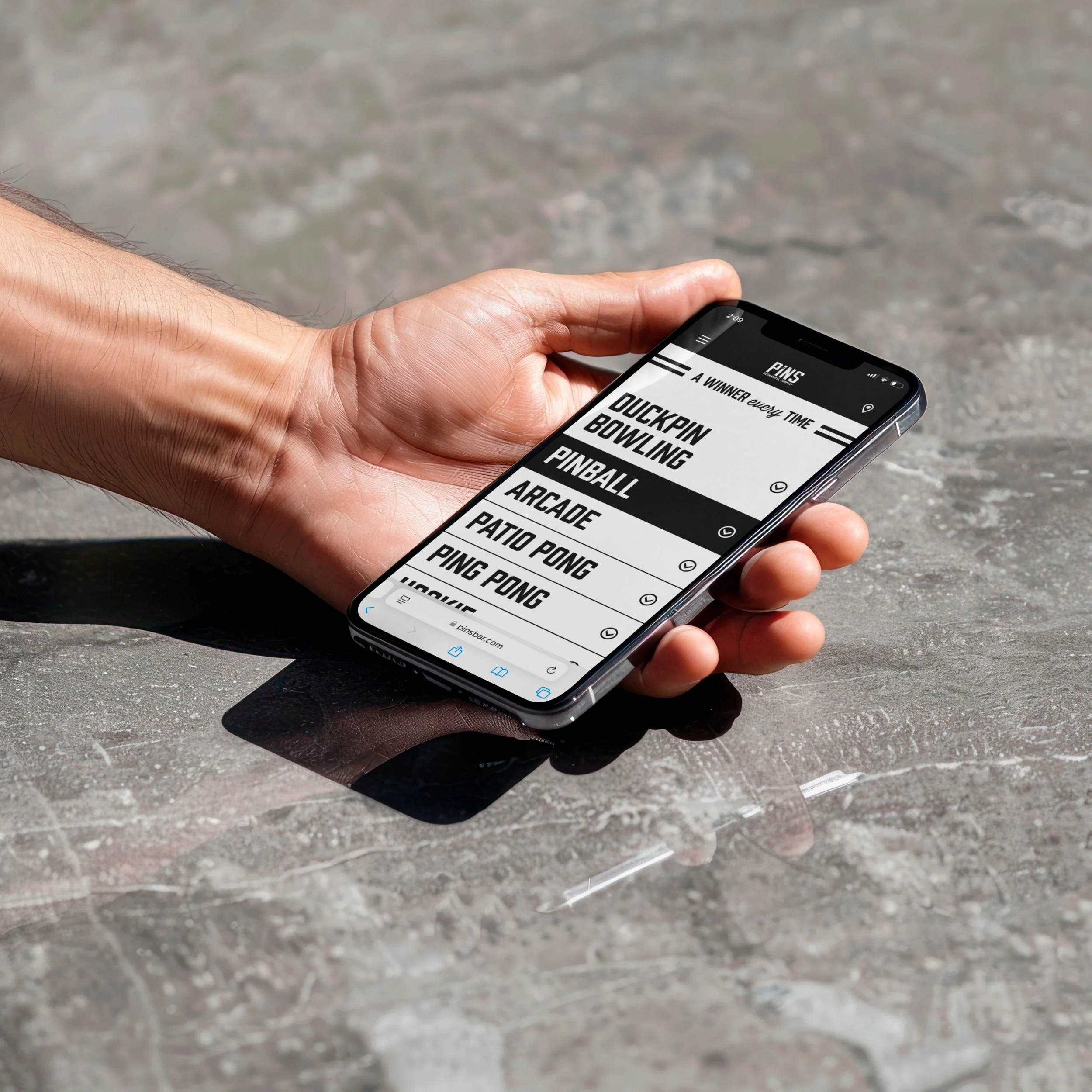 A person holding a smartphone with a digital pinboard app showing a list of arcade games, resting on a reflective stone surface outdoors.