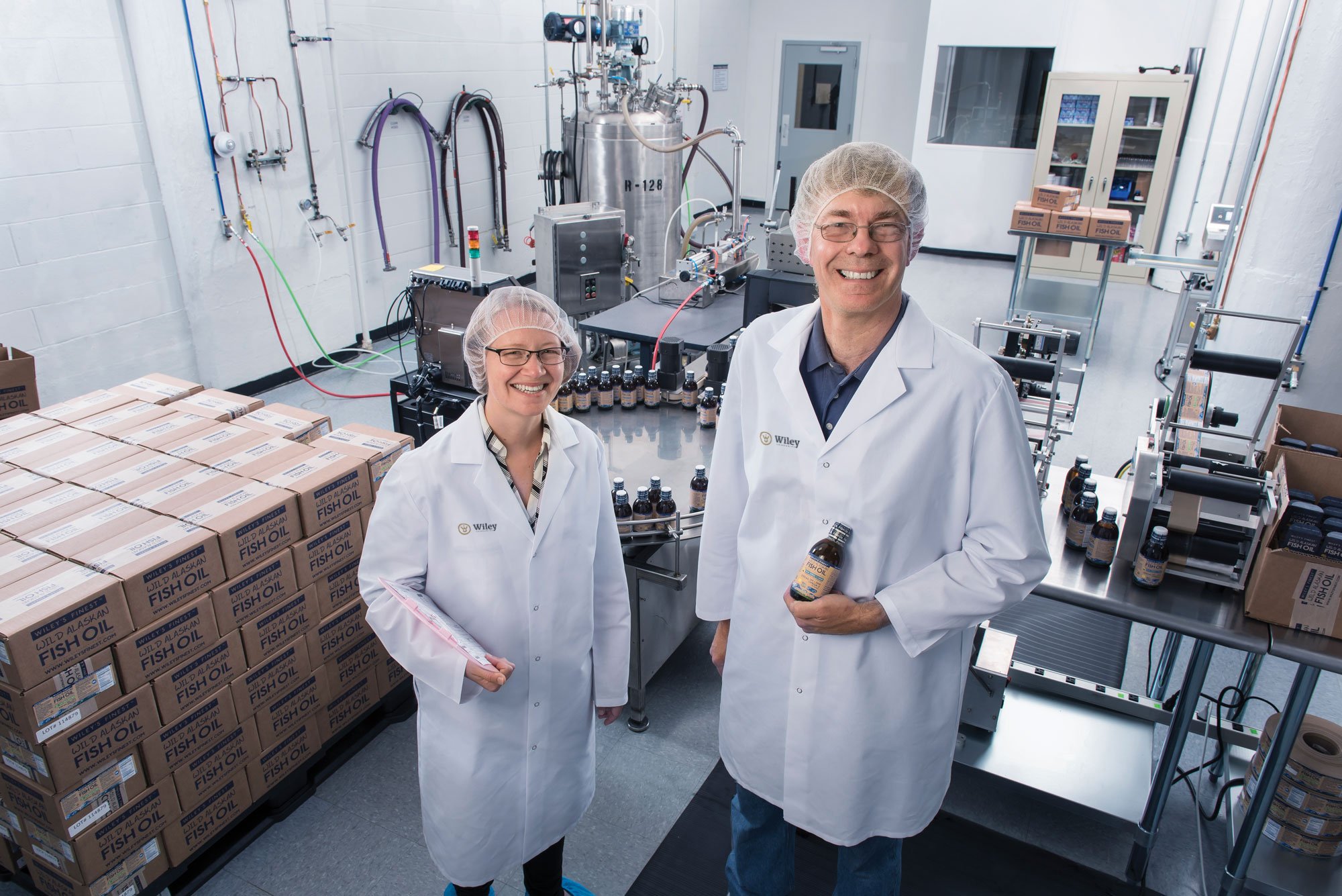 Two scientists in lab coats and hairnets in a laboratory, working with fish oil products. The woman is holding a clipboard, the man is holding a bottle of fish oil supplement. Lab equipment and boxes of fish oil are visible in the background.