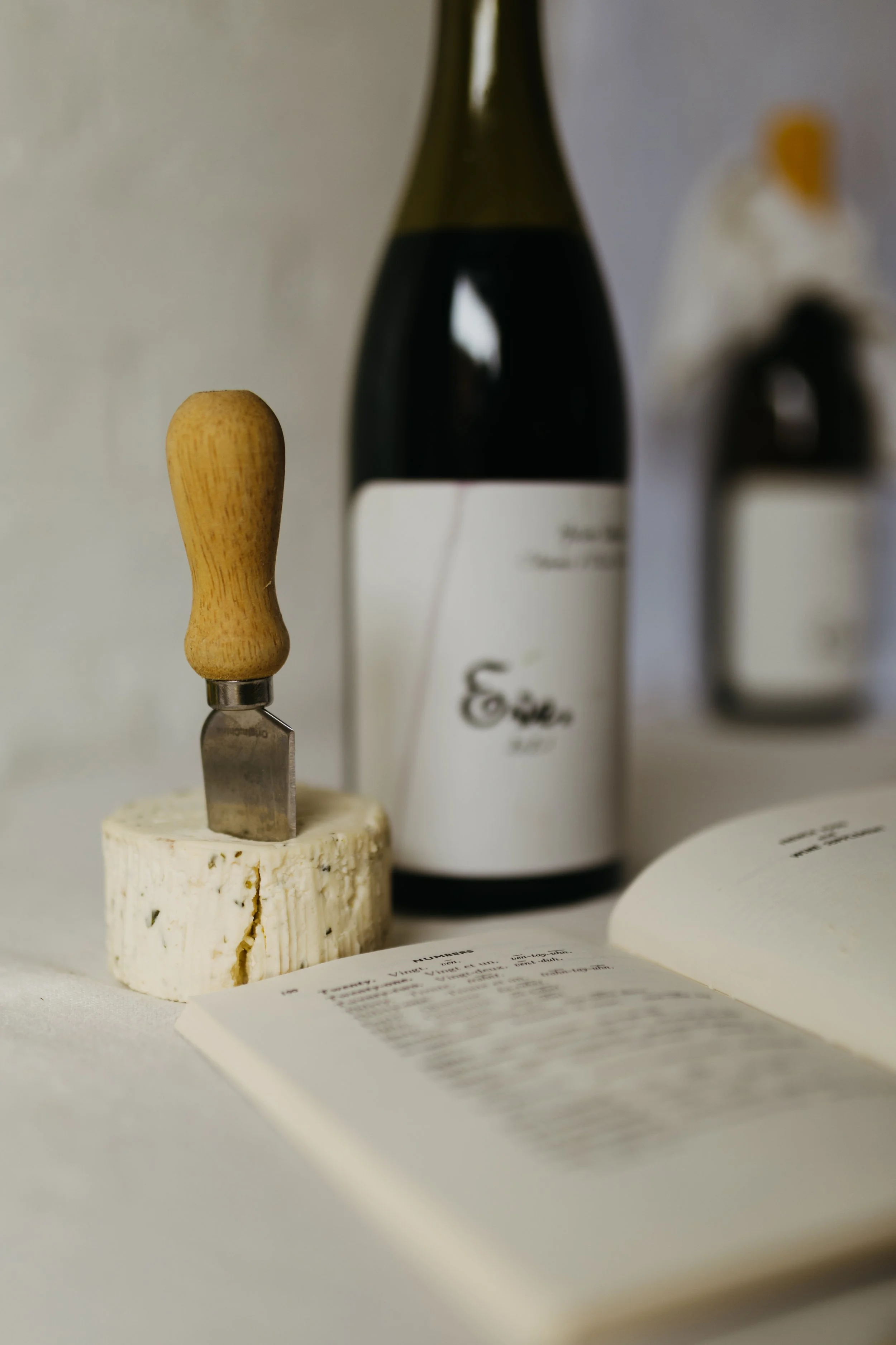 A small cheese knife with a wooden handle inserted into a round piece of cheese. Behind the cheese, there is a wine bottle with a white label. To the right, there is an open book and a blurred background with various objects.
