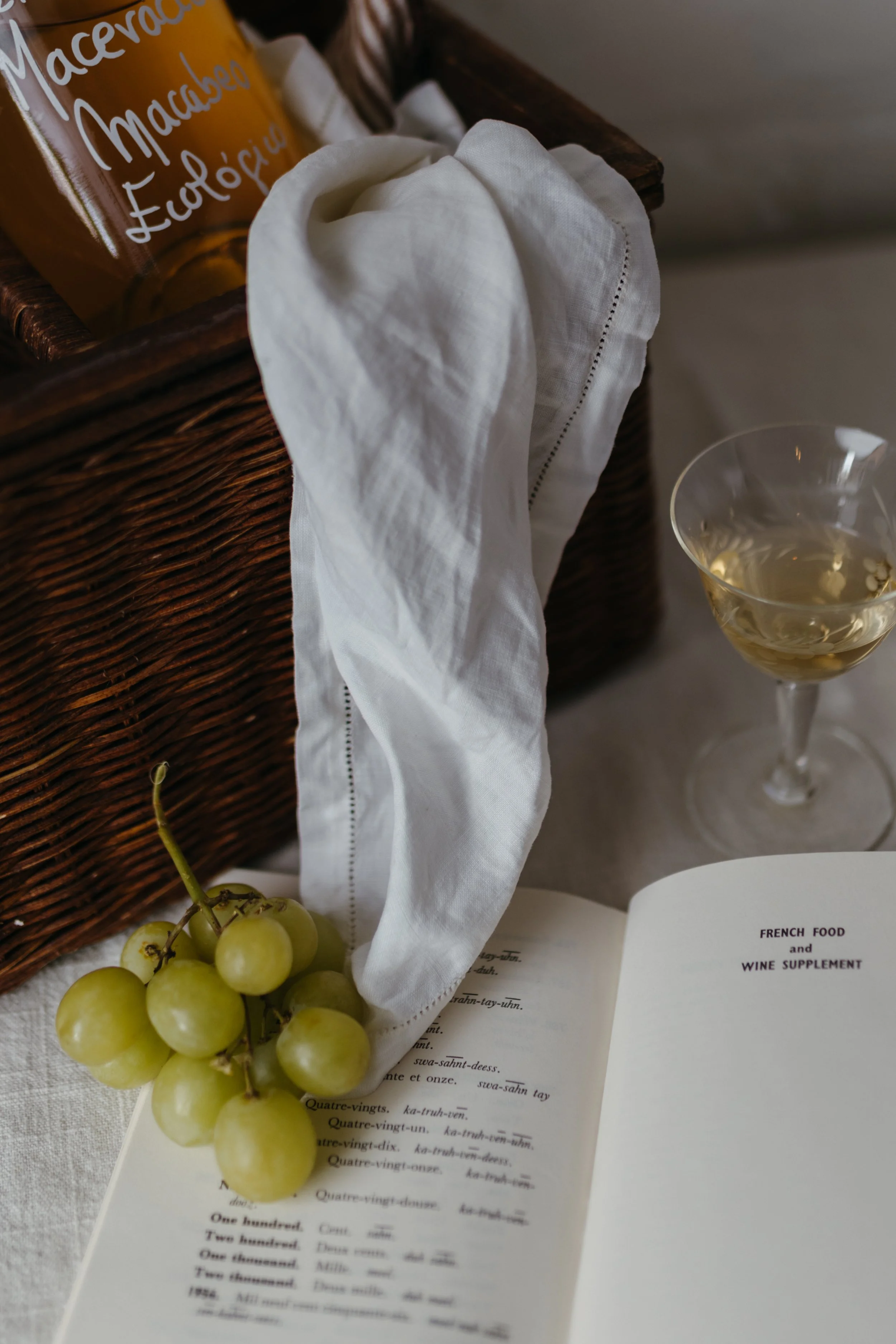 A bunch of green grapes, an open book with the title 'French Food and Wine Supplement', a white cloth hanging over a wicker basket, and a glass of white wine on a table.