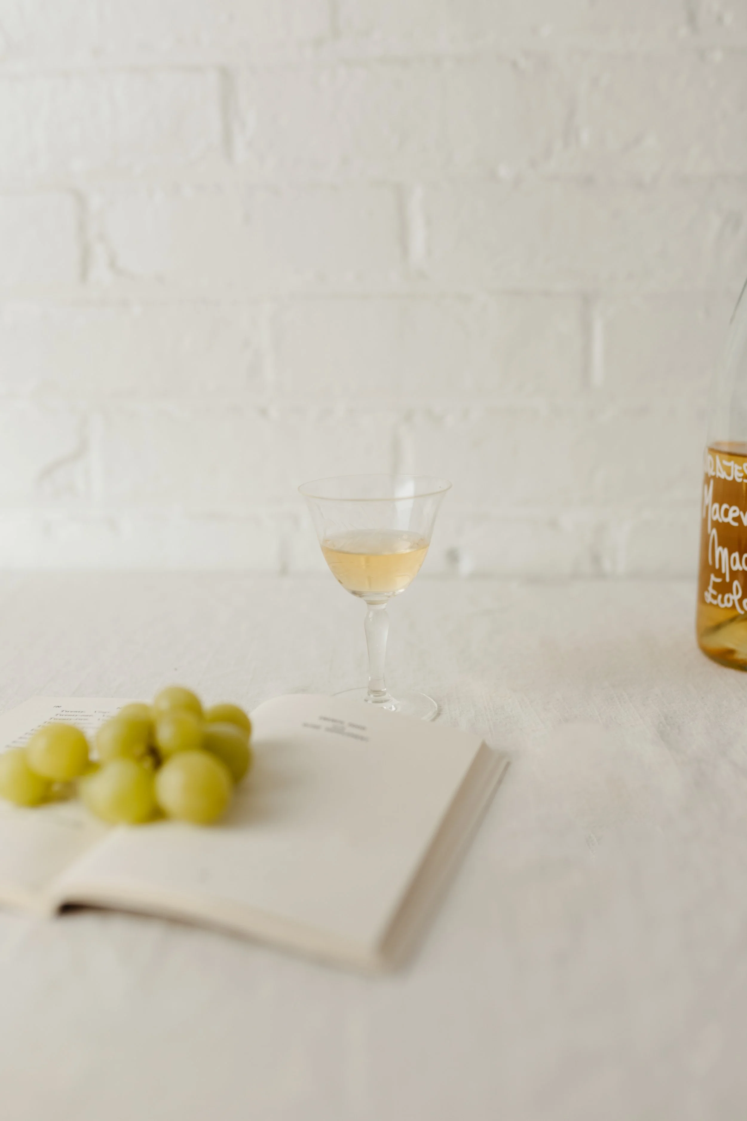 A glass of white wine on a white table next to a bunch of green grapes, an open book, and a bottle of wine.