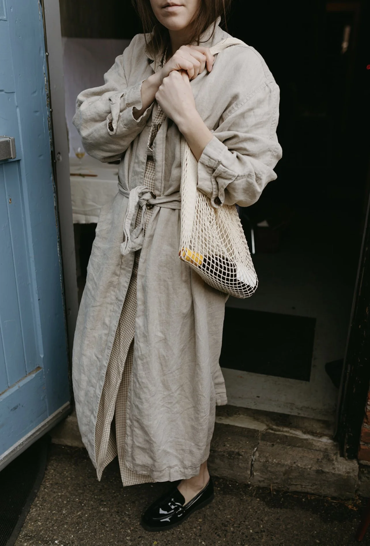 A woman standing outside a blue door holding a mesh bag across her shoulder, wearing a light-colored dress and black shoes.
