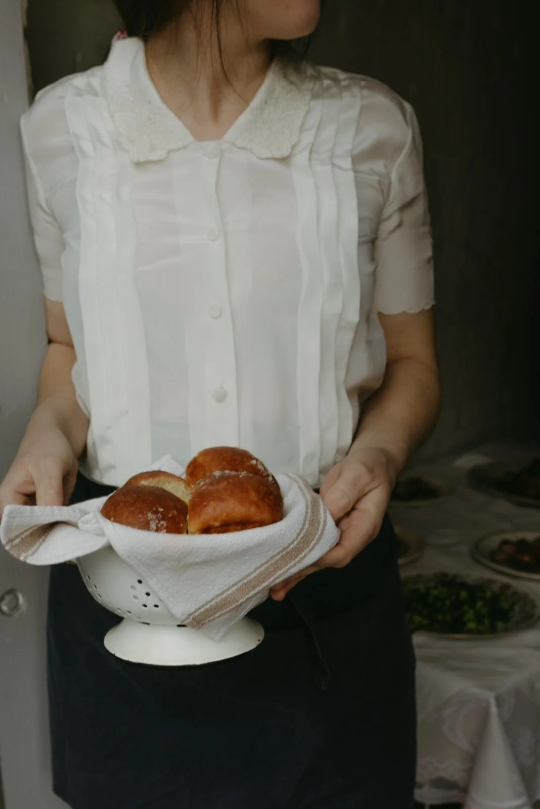 A woman holding a white colander with freshly baked bread rolls on a tea towel.