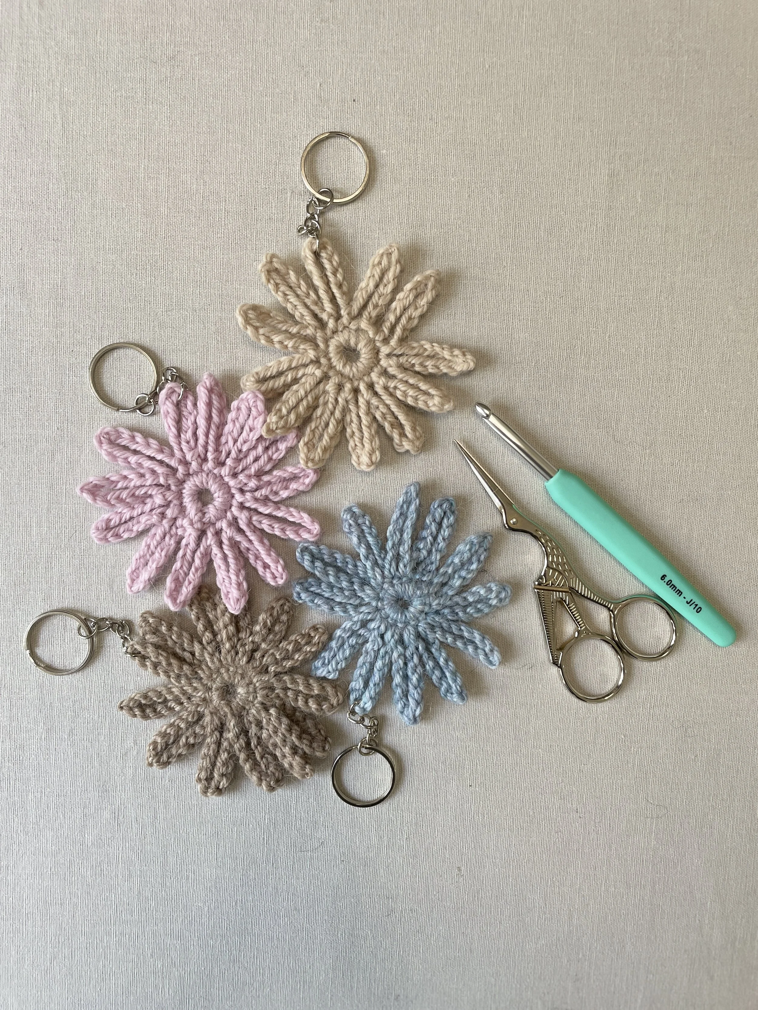 Three crocheted flower keychains in pink, beige, and blue, along with crochet scissors and a yarn needle on a light surface.