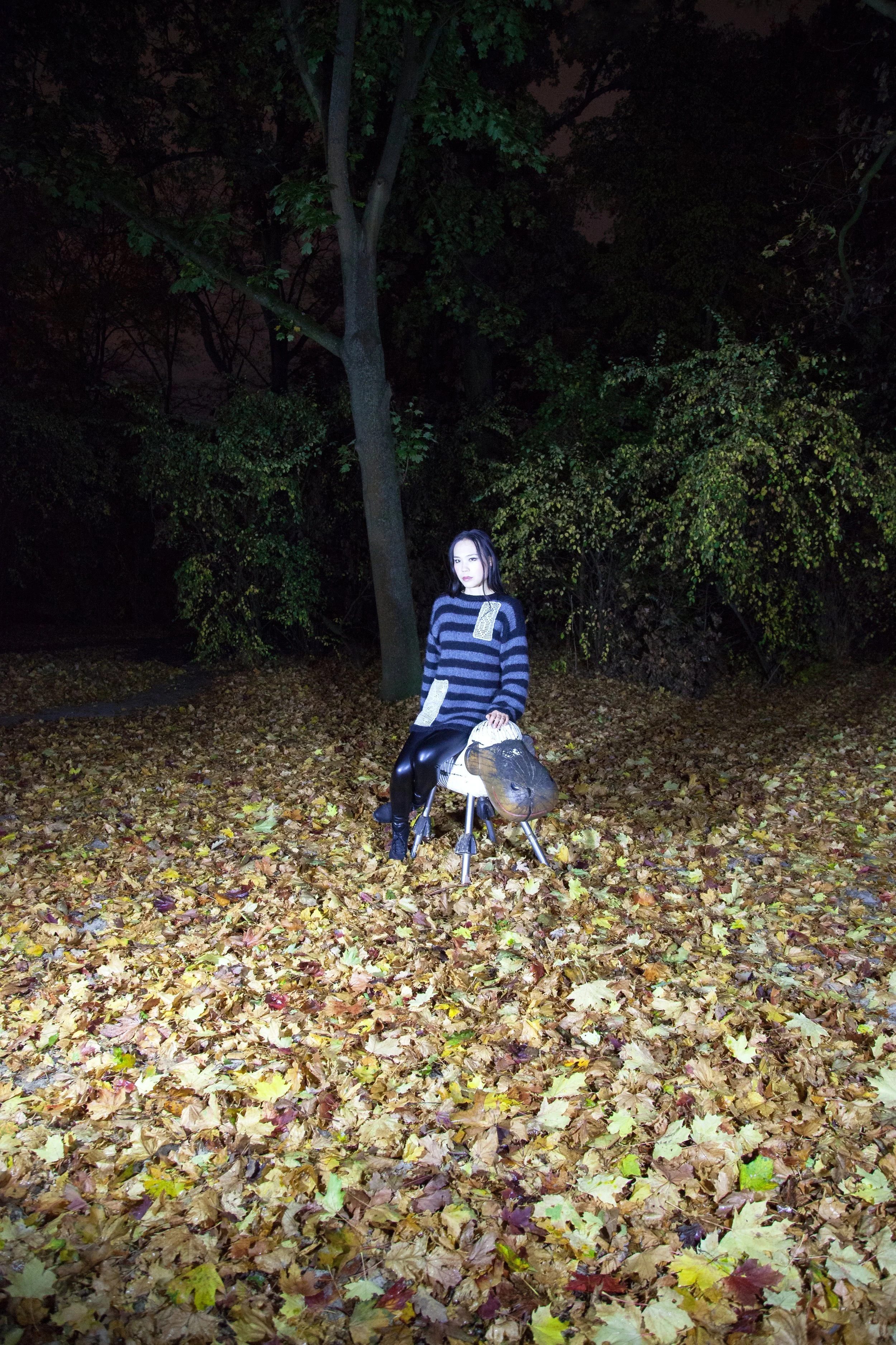 A woman sitting on a small, sheep sculpture in a forested area at night, surrounded by fallen autumn leaves.