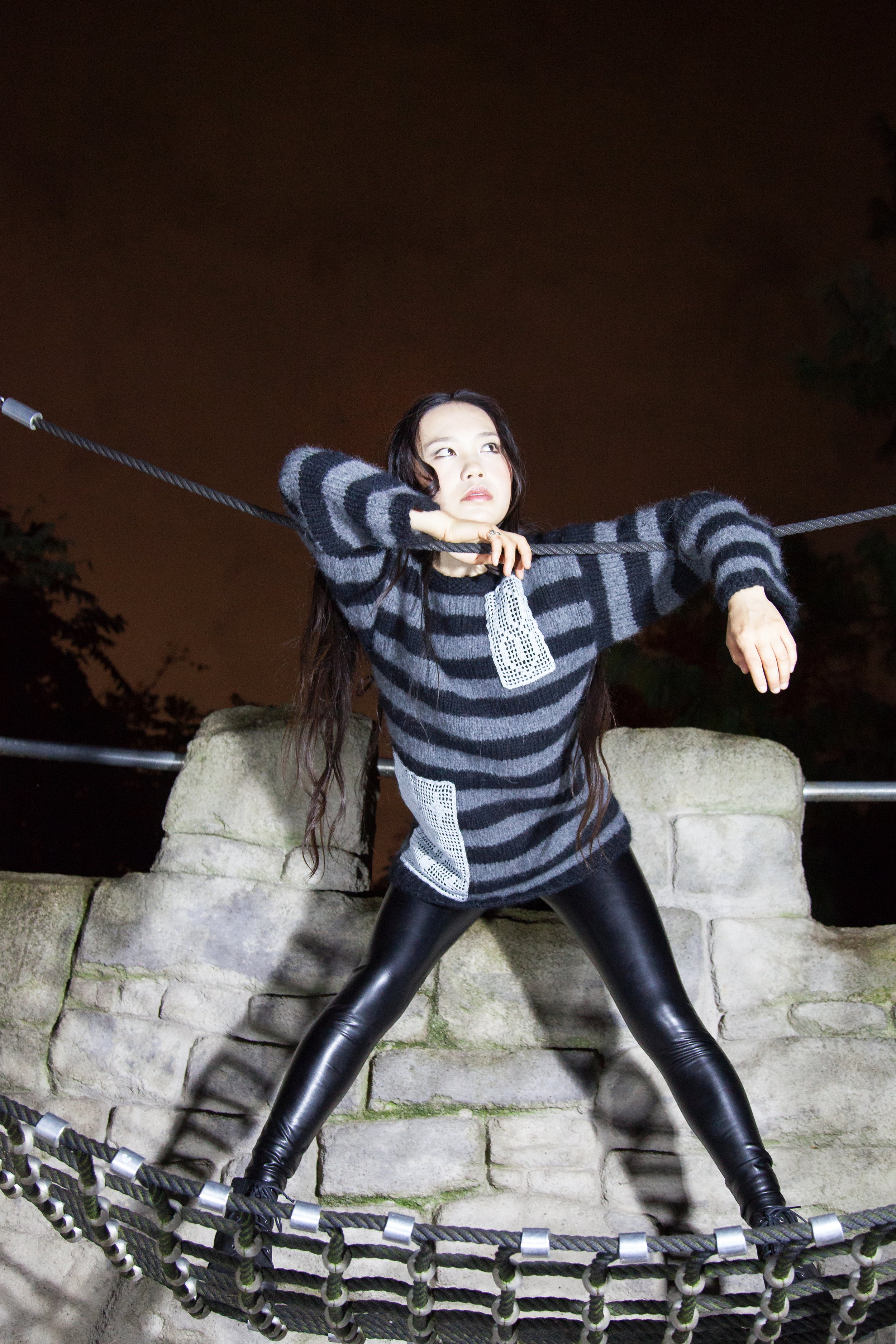 A woman with long dark hair wearing a black and gray striped sweater and black leather pants standing on a rope bridge at night, holding onto the ropes with both hands and looking upwards.