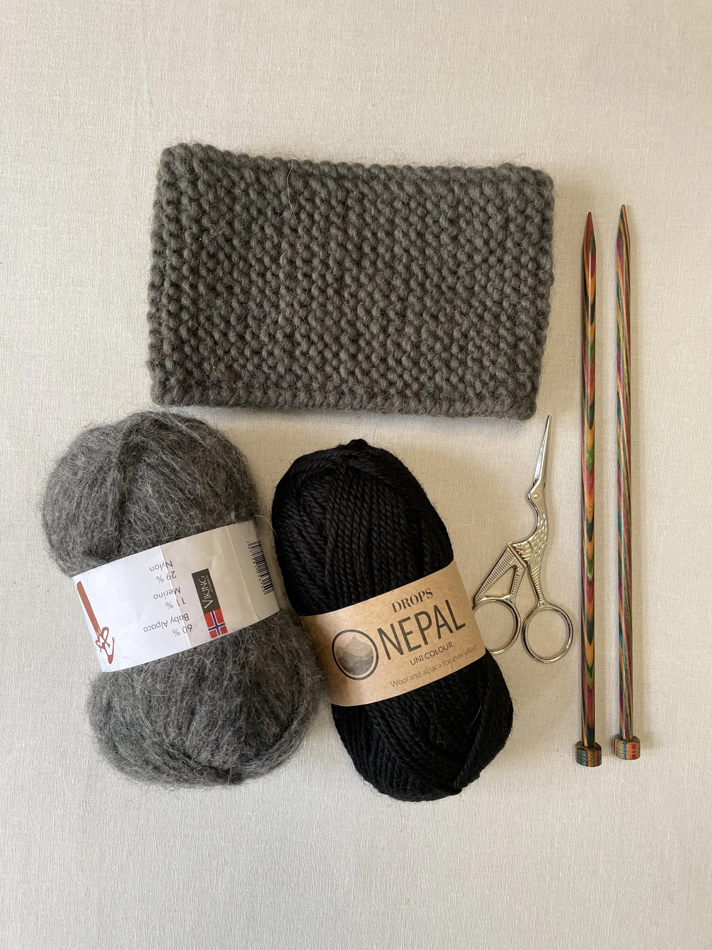 Two skeins of yarn, one gray and one black, a gray knitted headbans, a pair of small silver scissors with a curved tip, and two knitting needles with colorful patterns, arranged on a white surface.