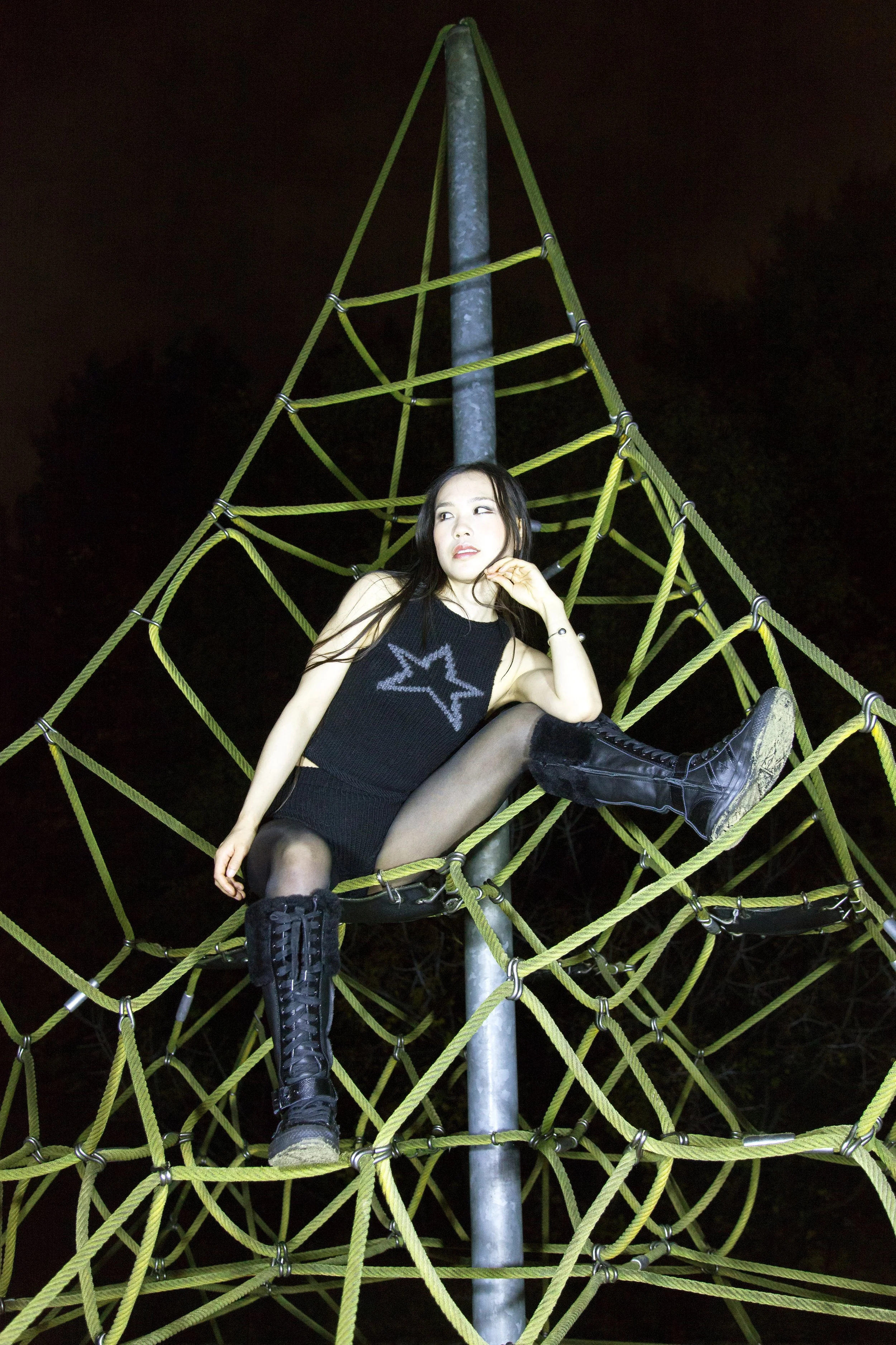 A woman sitting on a yellow rope playground climbing structure at night, wearing a black sleeveless top, black shorts, black tights, and black combat boots.